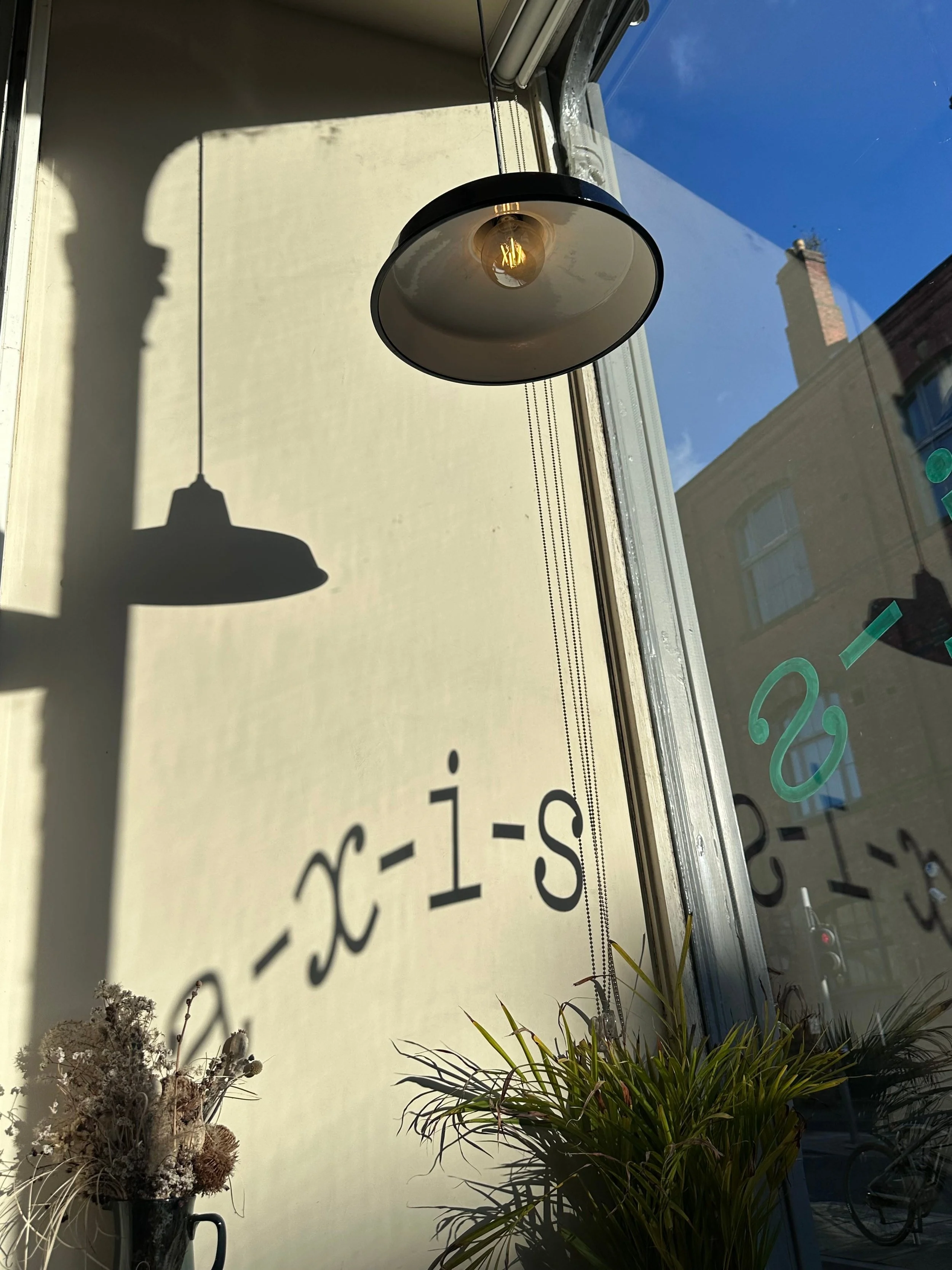 Hanging lamp casting shadow on sunlit wall with the word "axis" partially visible; green plants in foreground, clear blue sky outside. Calm ambiance.