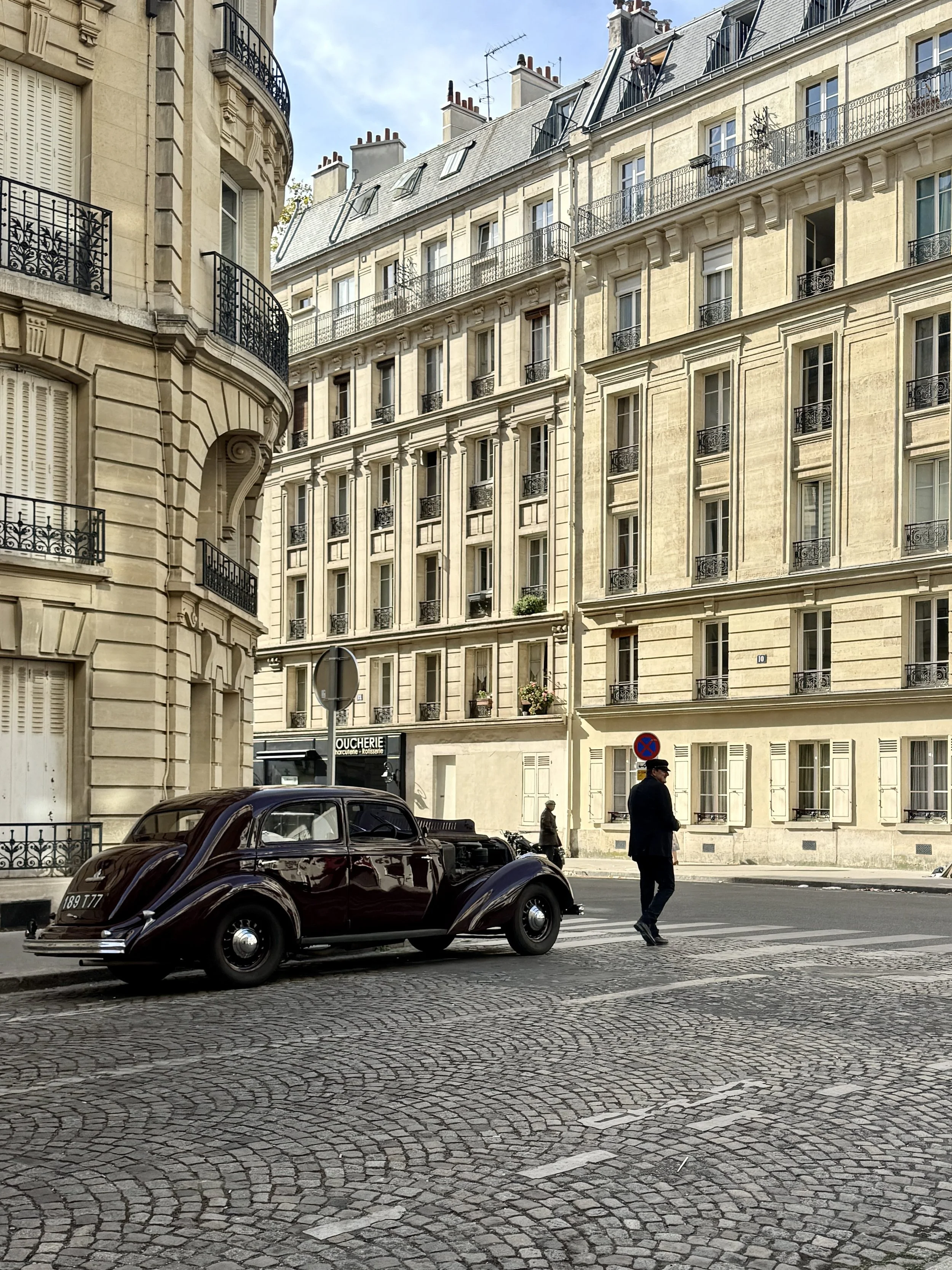 A glossy vintage black car crosses a cobblestone street beside elegant Parisian stone buildings, as a lone pedestrian passes in bright sunlight.