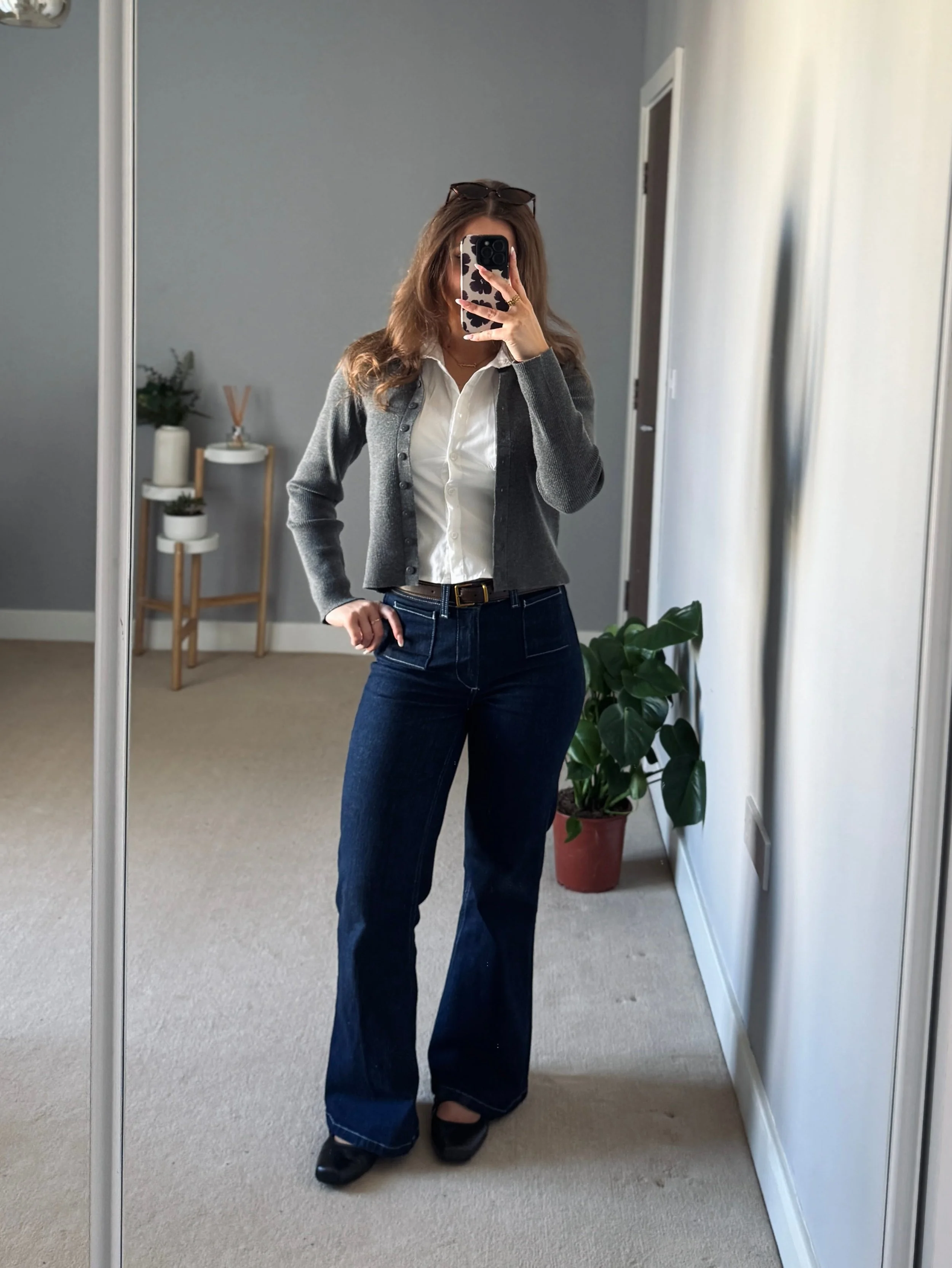 Woman taking a mirror selfie in a minimalist room. She wears flared jeans, white shirt, gray cardigan. A plant and shelf add decor. Casual mood.
