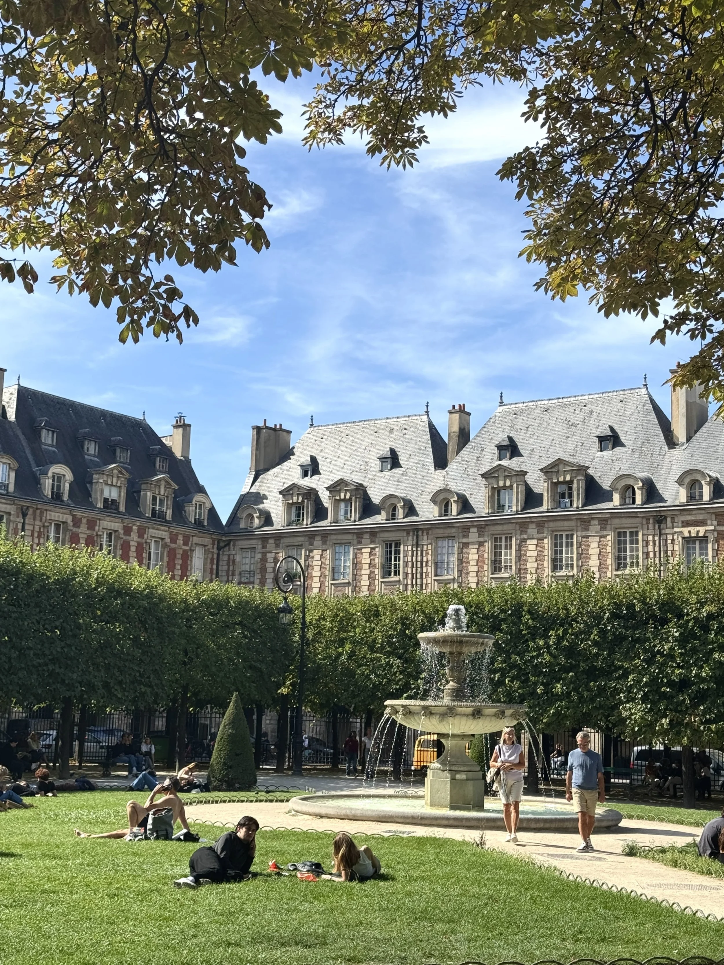 Sunlit Parisian courtyard with a stone fountain, clipped trees, and historic mansard-roofed buildings; visitors relax on the lawn in a peaceful scene.