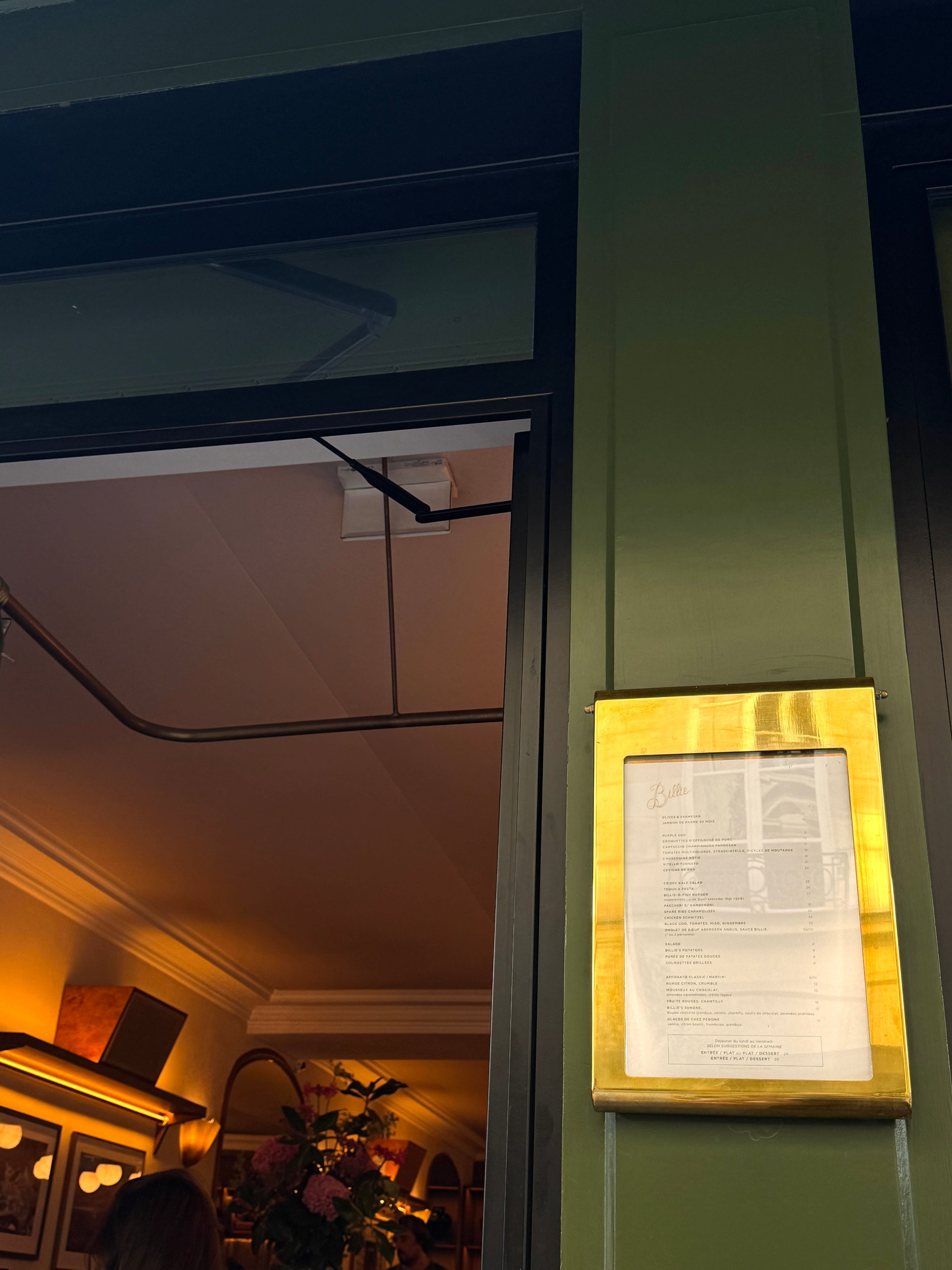 A glowing yellow-framed menu hangs on a dark green wall beside an open doorway, revealing a warmly lit room with flowers and lamps.
