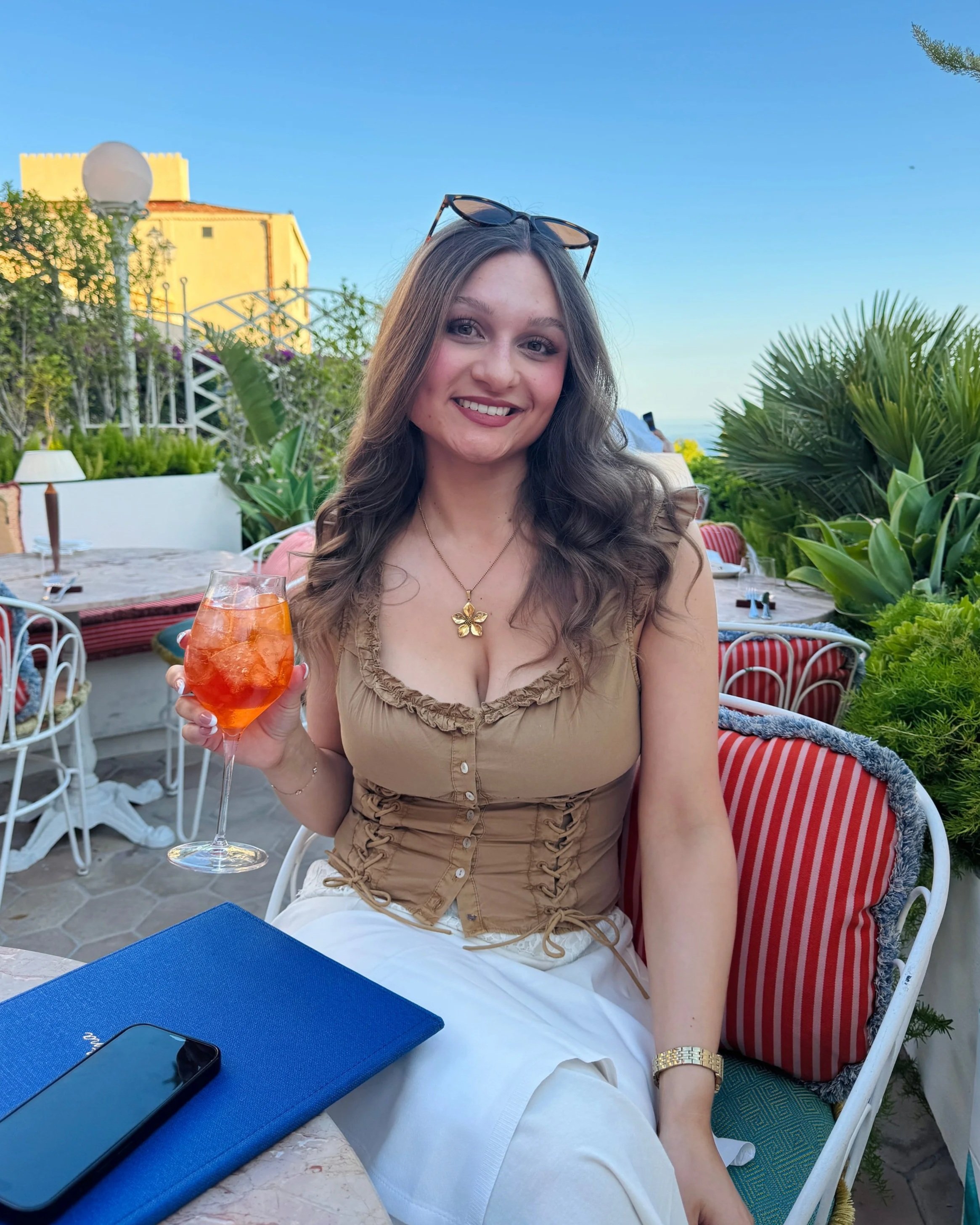 A woman sitting at an outdoor table in a cafe, holding a drink, wearing a tan fitted top and white pants, with plants and blue sky in the background.