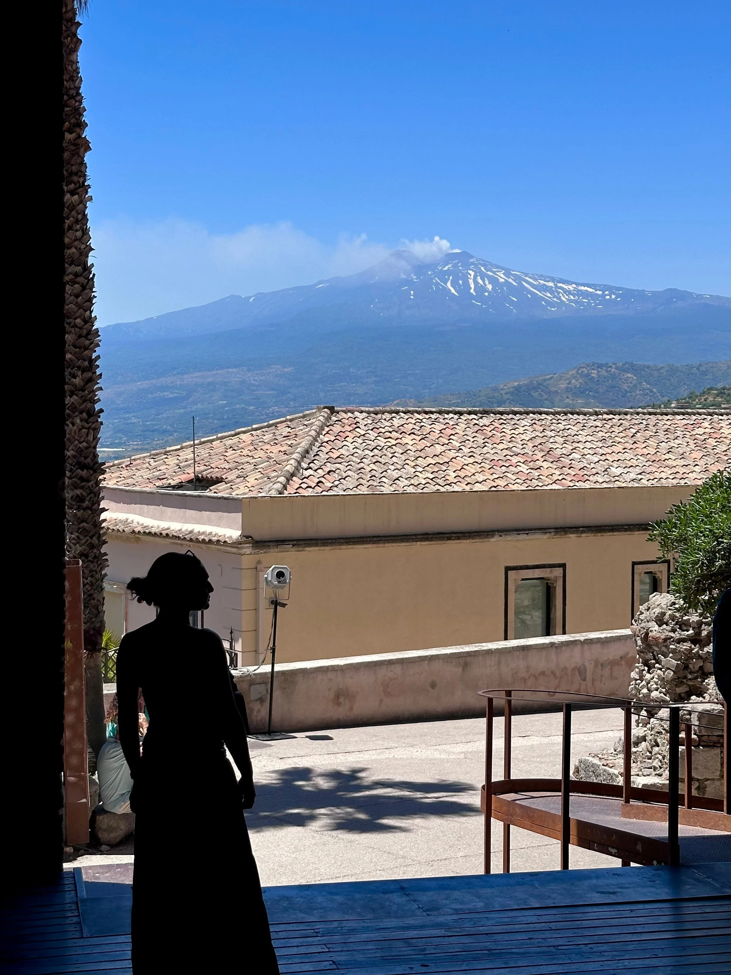 Silhouette of a person standing in a doorway overlooking rooftops, with a smoking snow-capped mountain and clear blue sky in the background.