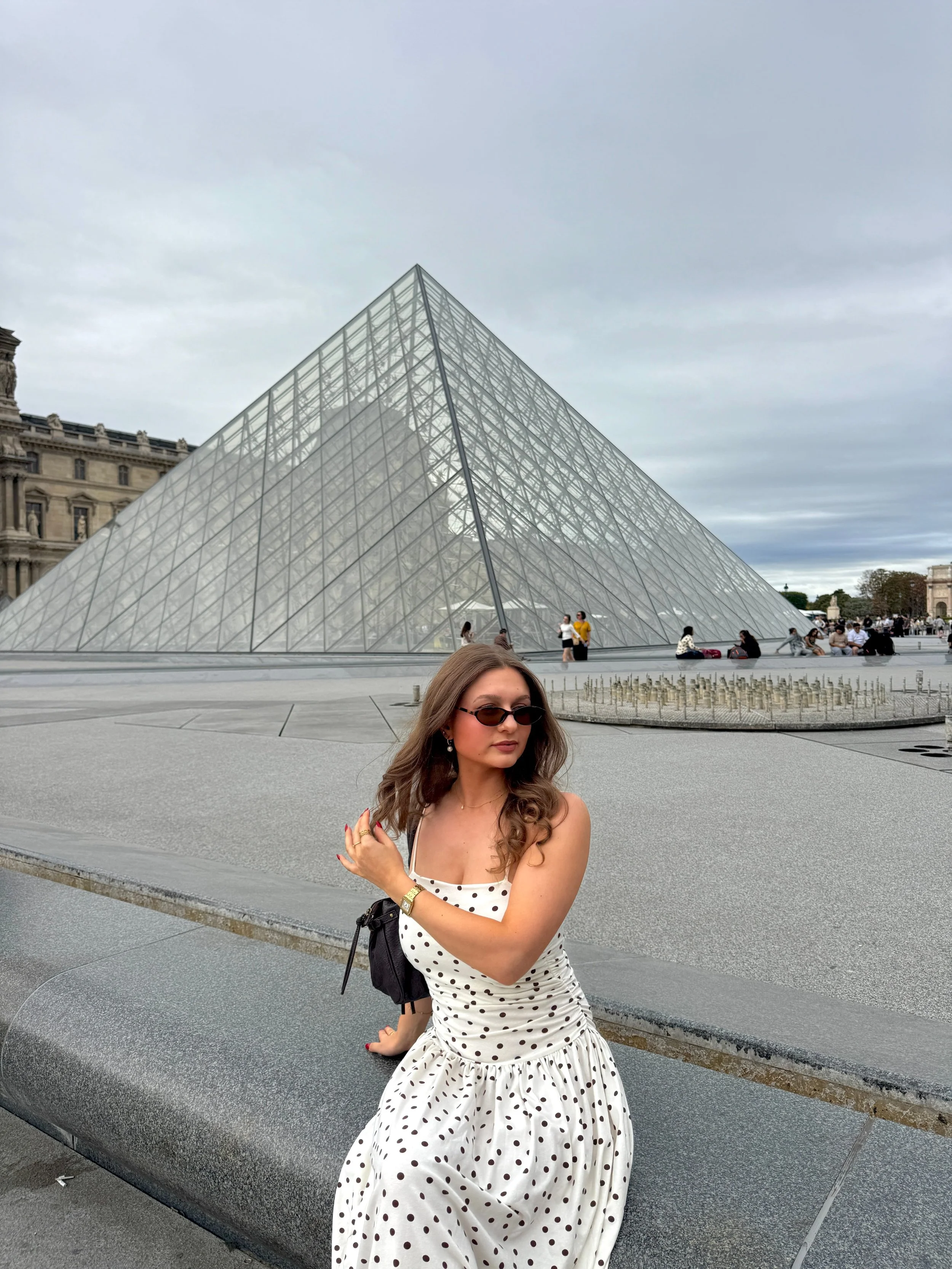Woman in a white polka-dot dress and sunglasses poses before the Louvre Pyramid in Paris under an overcast sky, creating a stylish travel scene.
