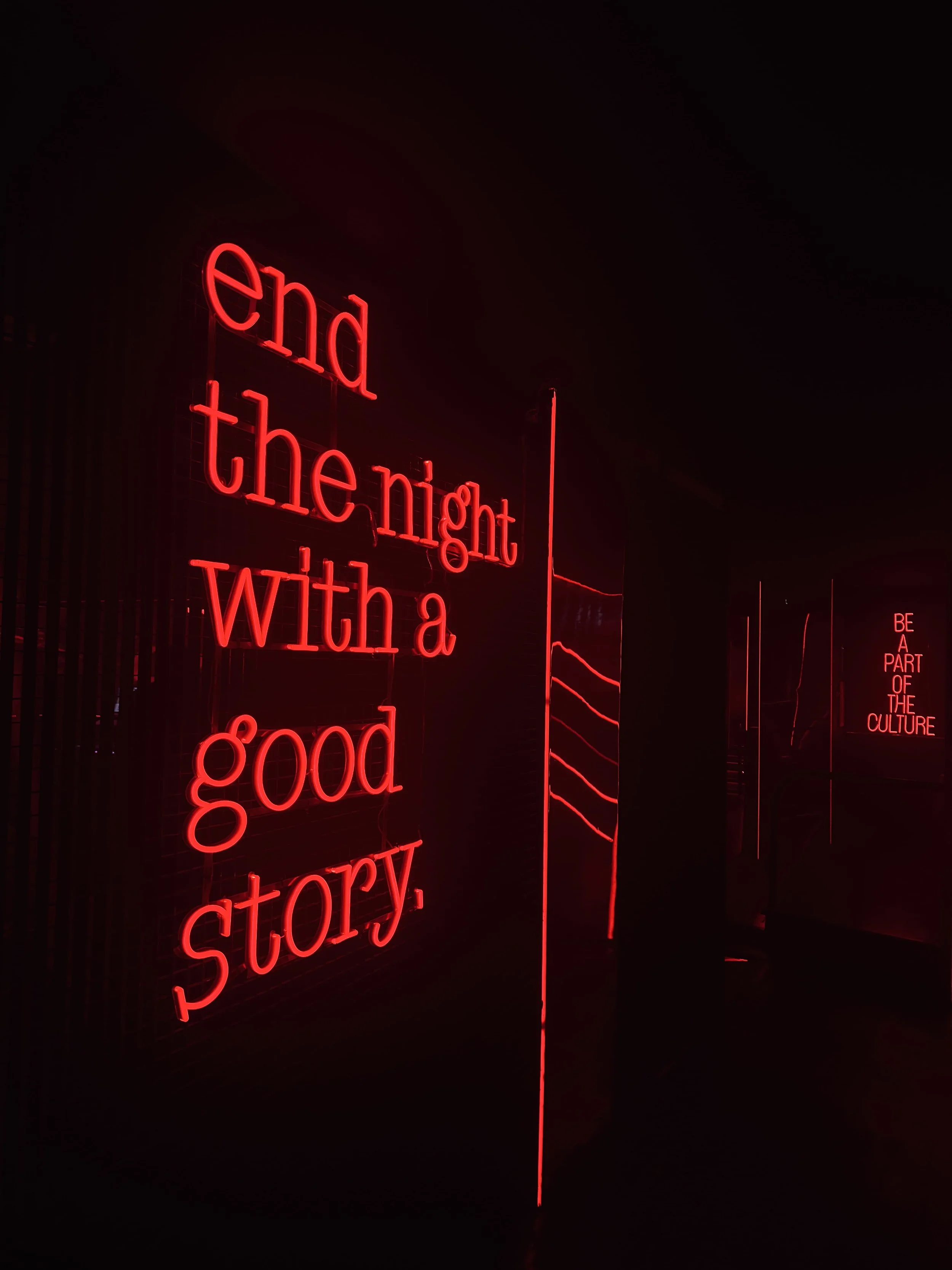 Red neon text reading “end the night with a good story” glows on a dark wall, with a smaller “Be a part of the culture” sign in the distance. An atmospheric installation.