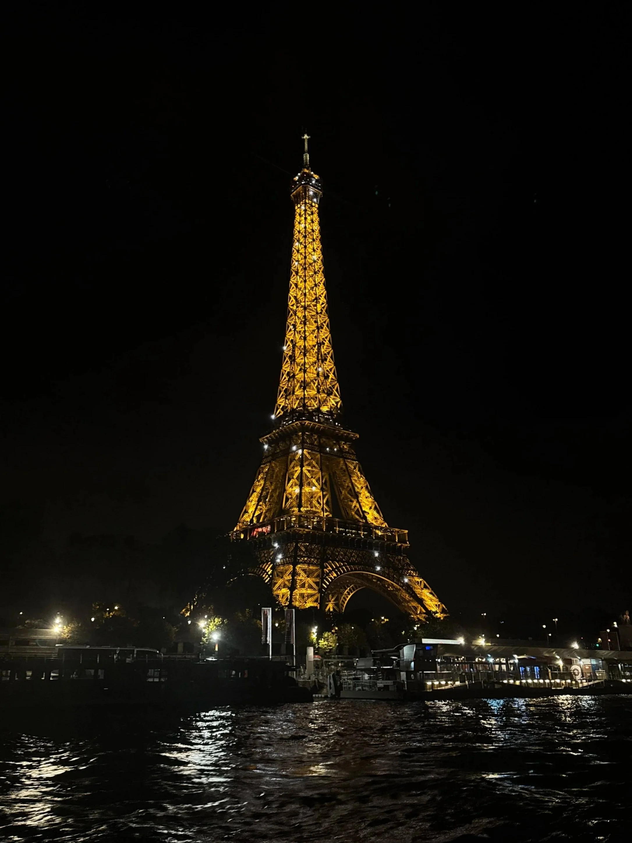 The Eiffel Tower glows golden against a black Paris night, viewed from a boat on the rippling Seine; the scene feels calm and romantic.