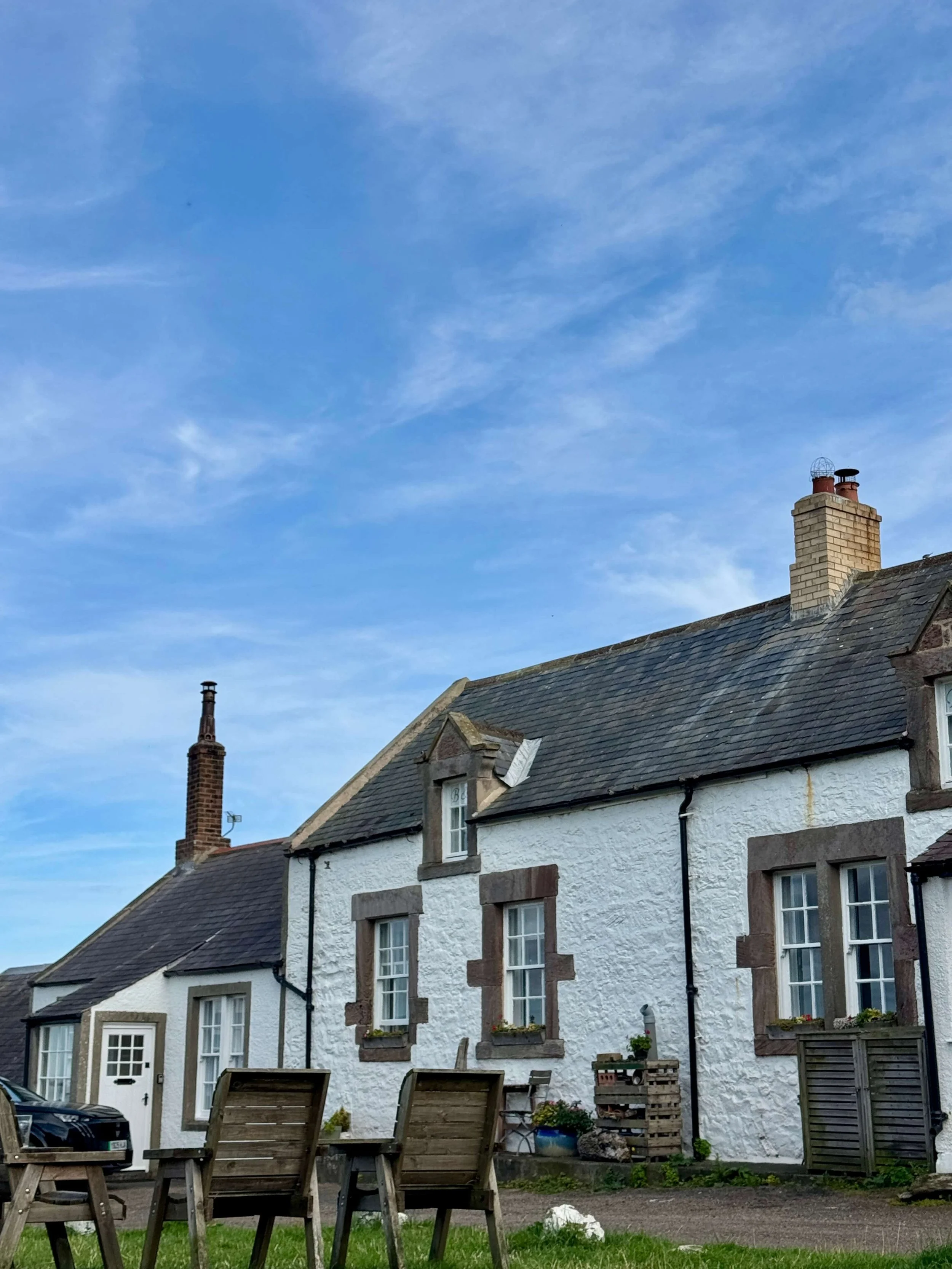 Whitewashed stone cottage with slate roofs, chimneys, and blue-sky backdrop; rustic chairs and potted plants create a peaceful village scene.