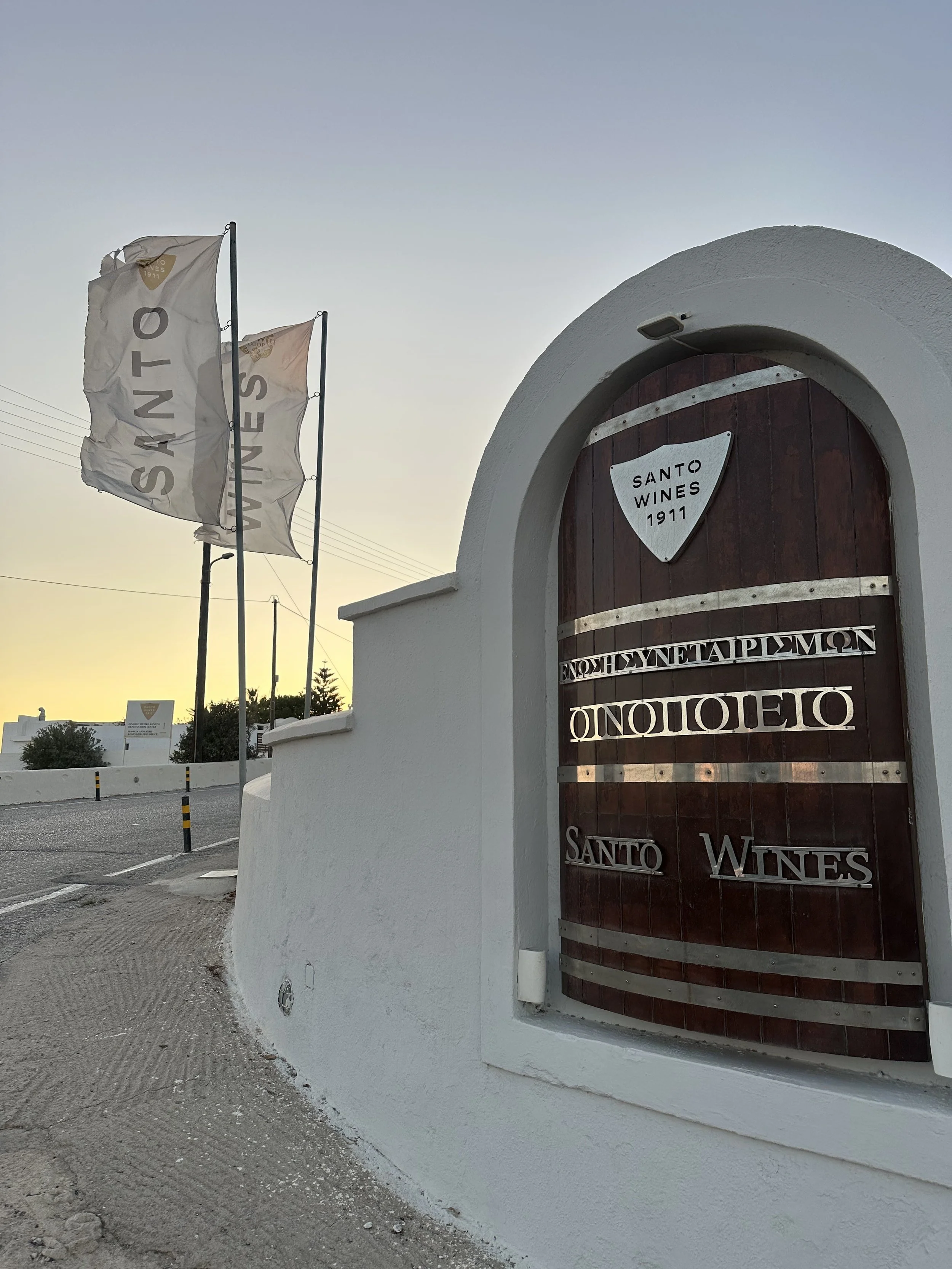 A barrel embellelished with a sign saying 'Santo Wines' at the entrance of the Santo Wines winery in Santorini, with two flags in the background.