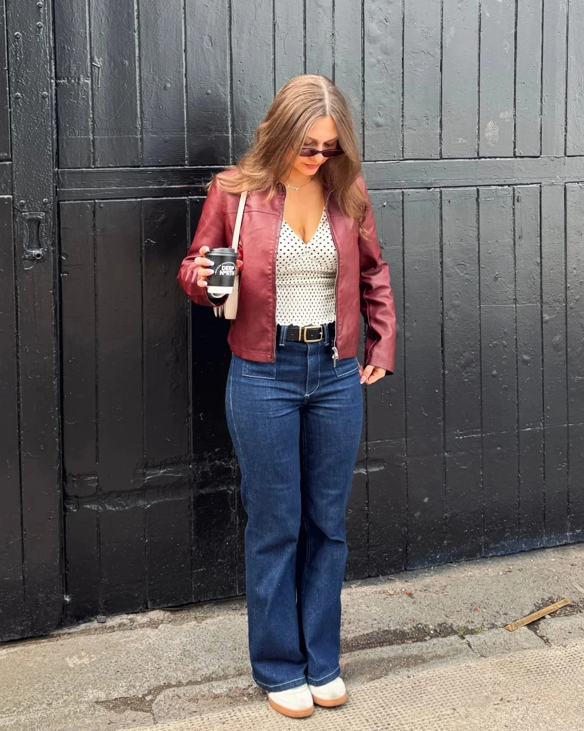 A woman stands in front of a black wooden door, wearing a maroon jacket, polka dot top, blue jeans, and white shoes. She holds a coffee cup, and is looking down.