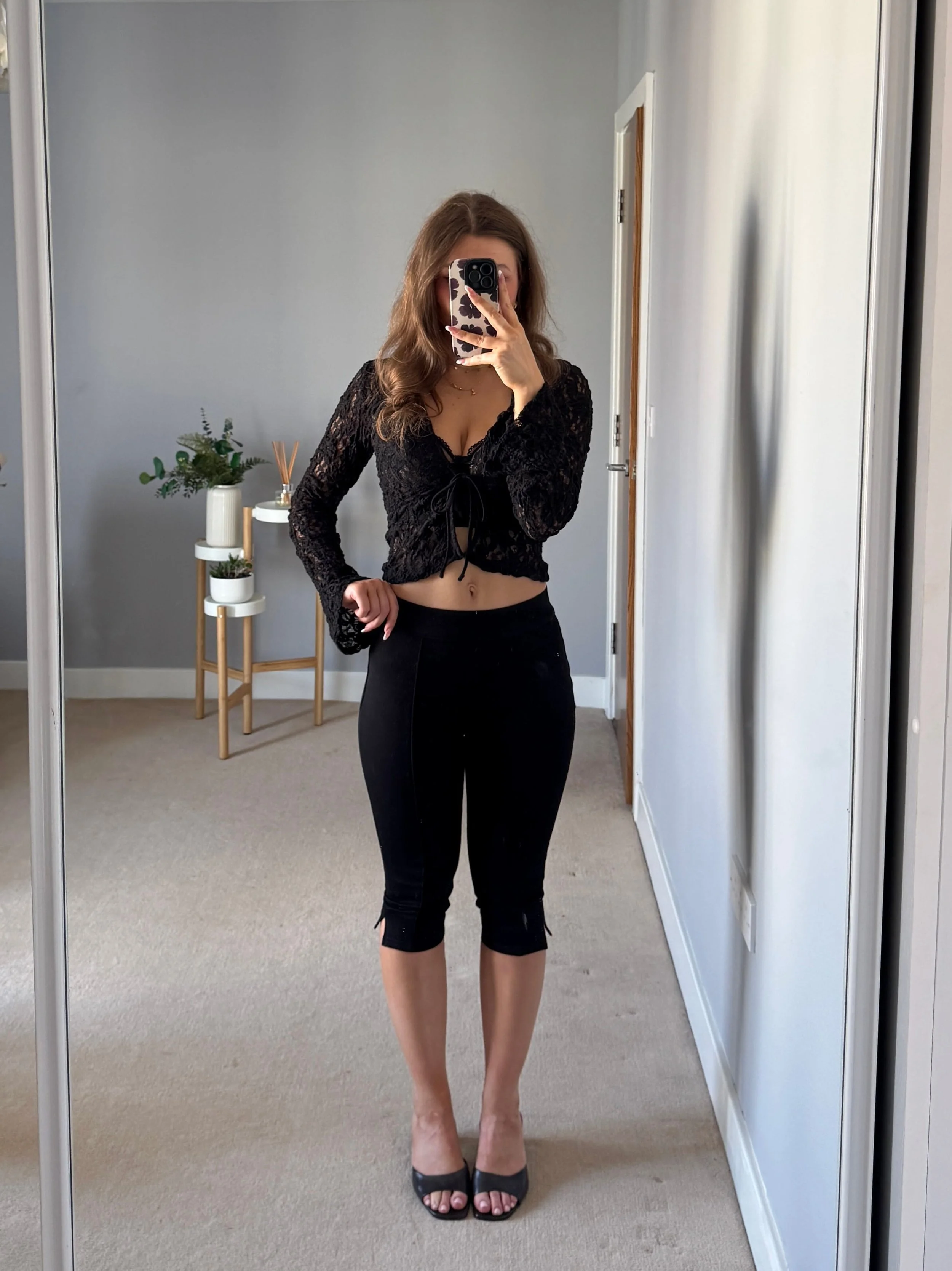 Woman taking mirror selfie wearing black lace crop top, black capri leggings, and black open-toe heels in a minimalist room with light grey walls.