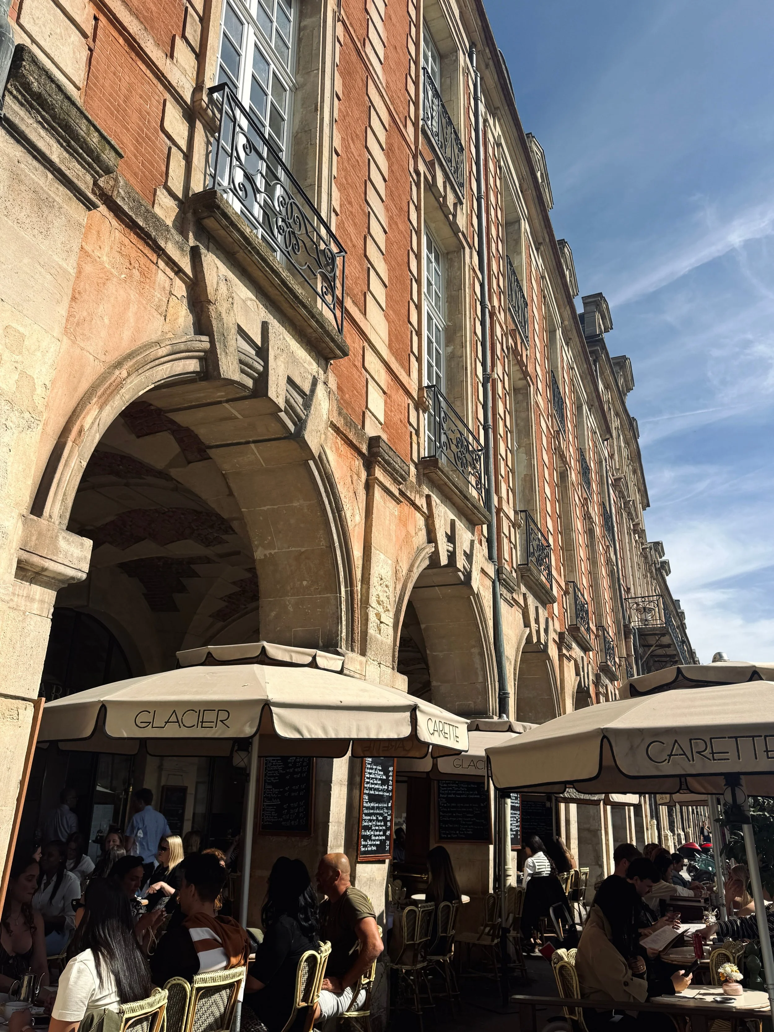 Low-angle view of a sunlit red-brick European building with stone arches and tall windows; café terraces bustle beneath beige umbrellas, creating a lively, historic street scene.