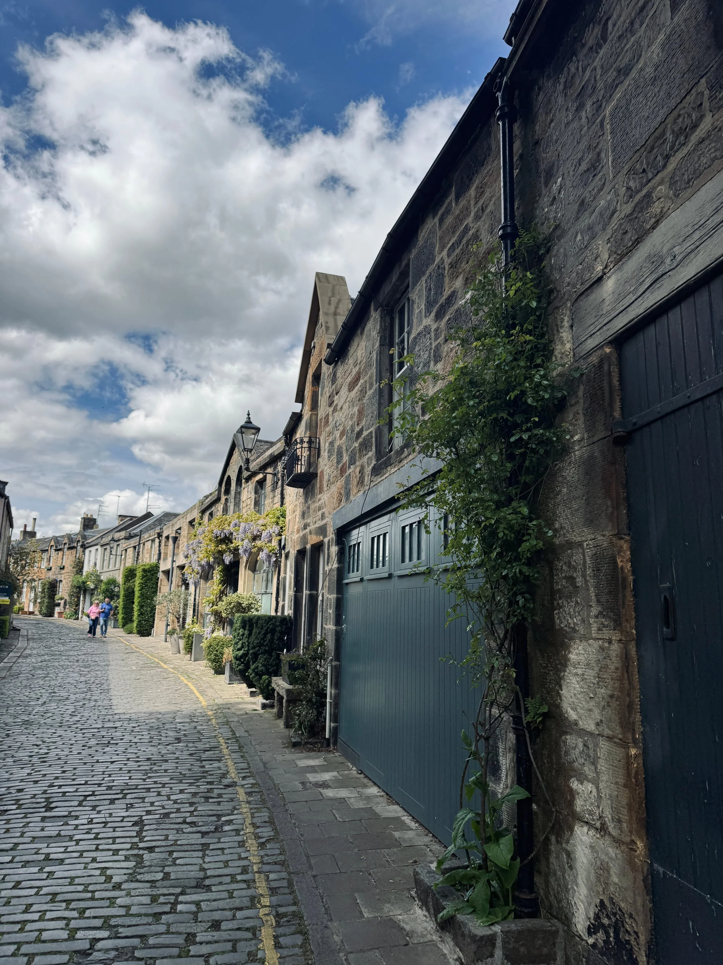 A narrow, cobblestone street lined with quaint stone buildings under a partly cloudy sky. Ivy climbs a wall, and a couple walks in the distance.