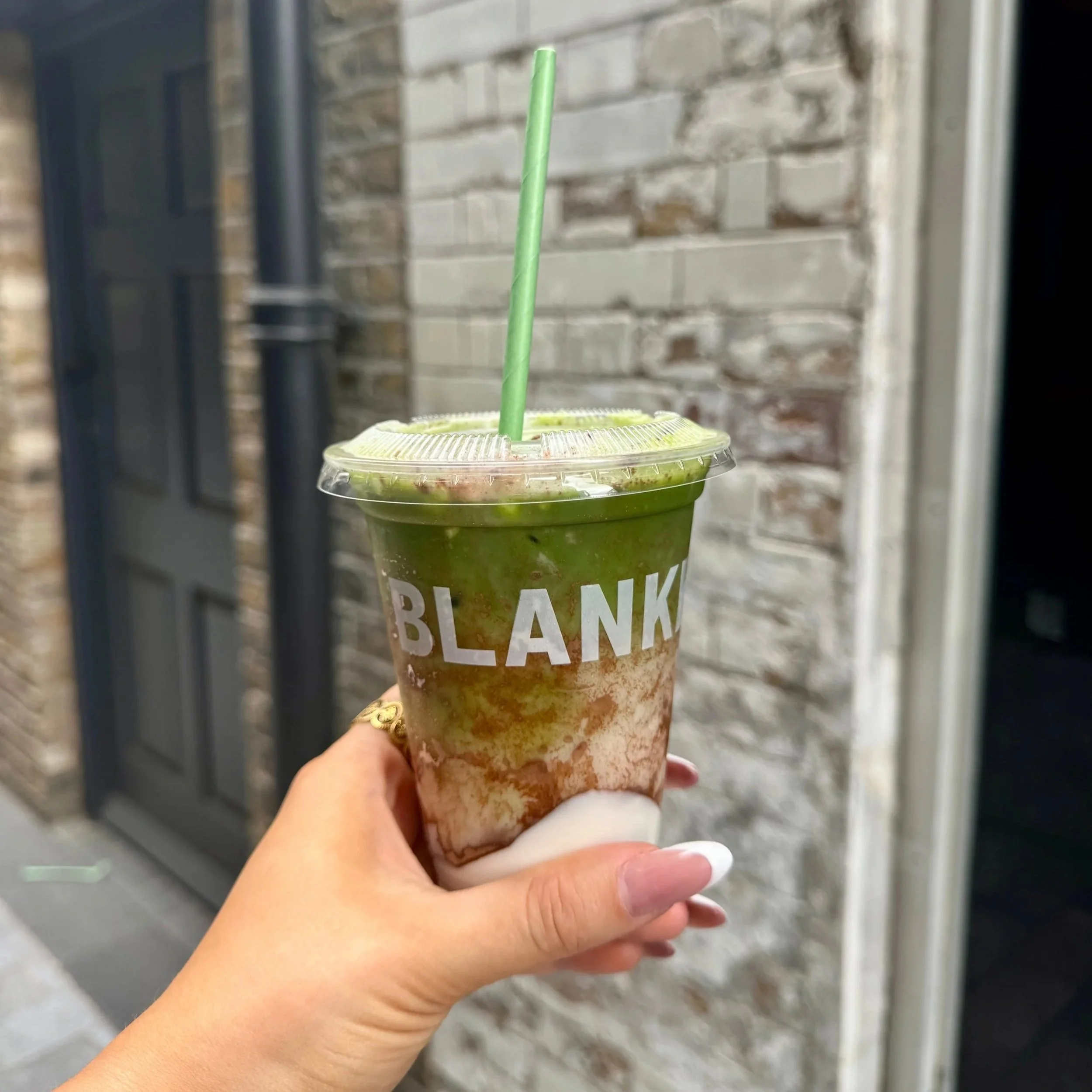 A person holds a vibrant green matcha drink in front of a rustic brick building.
