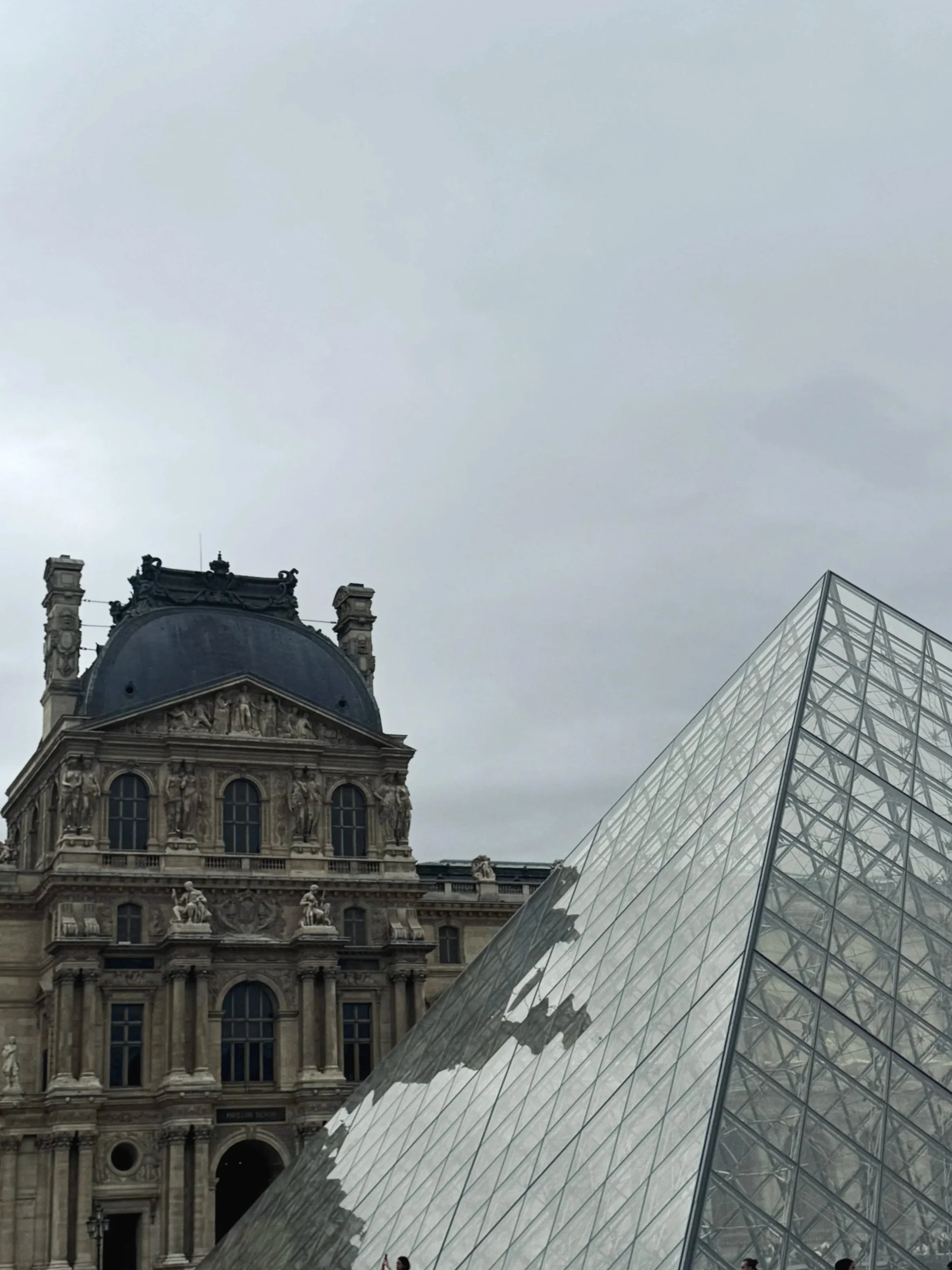 The Louvre’s glass pyramid rises beside the historic palace under a gray sky, with visitors gathered at its base; the scene feels grand and subdued.