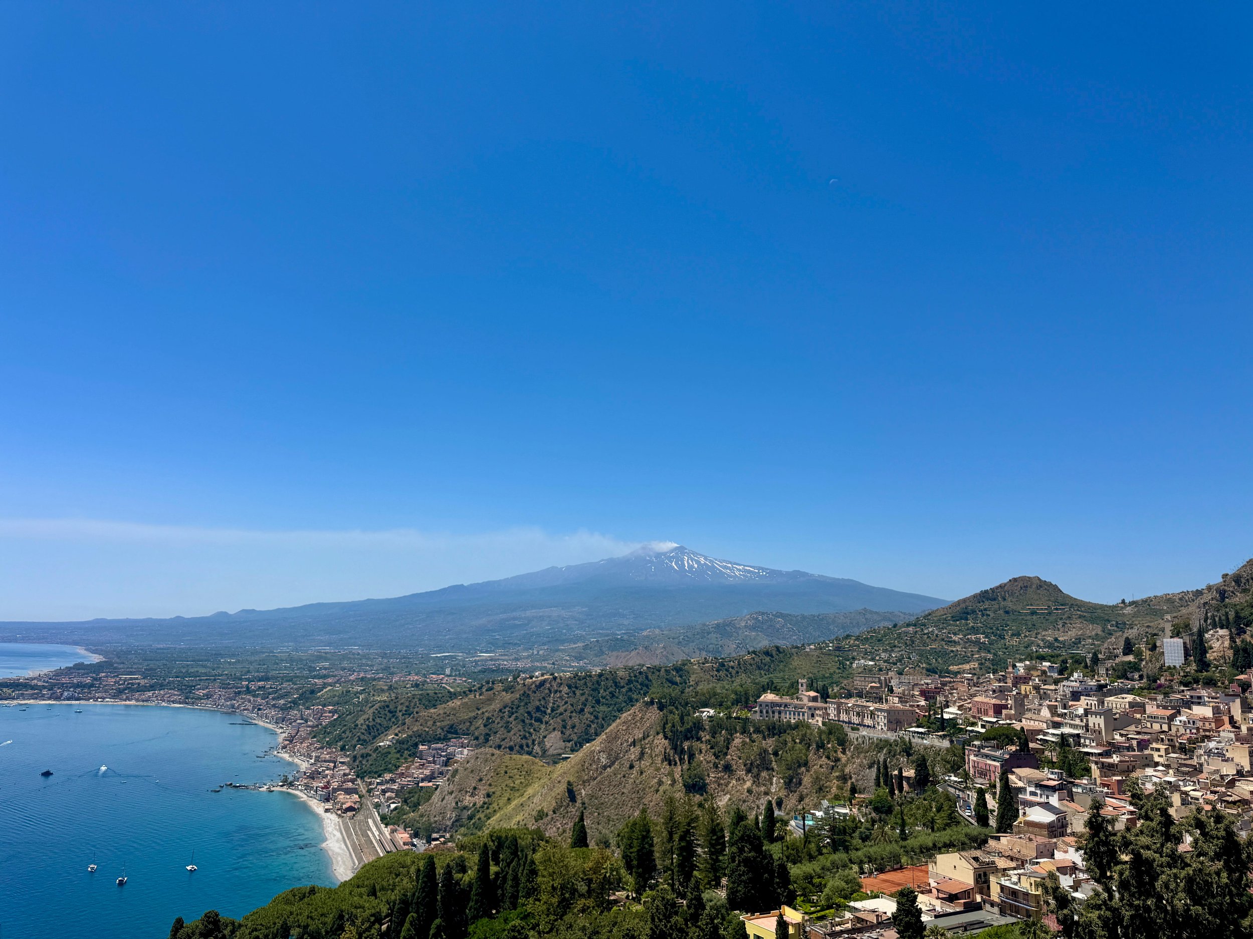 Panoramic coastal town view with blue sea, lush hills, and snow-capped mountain under clear sky.