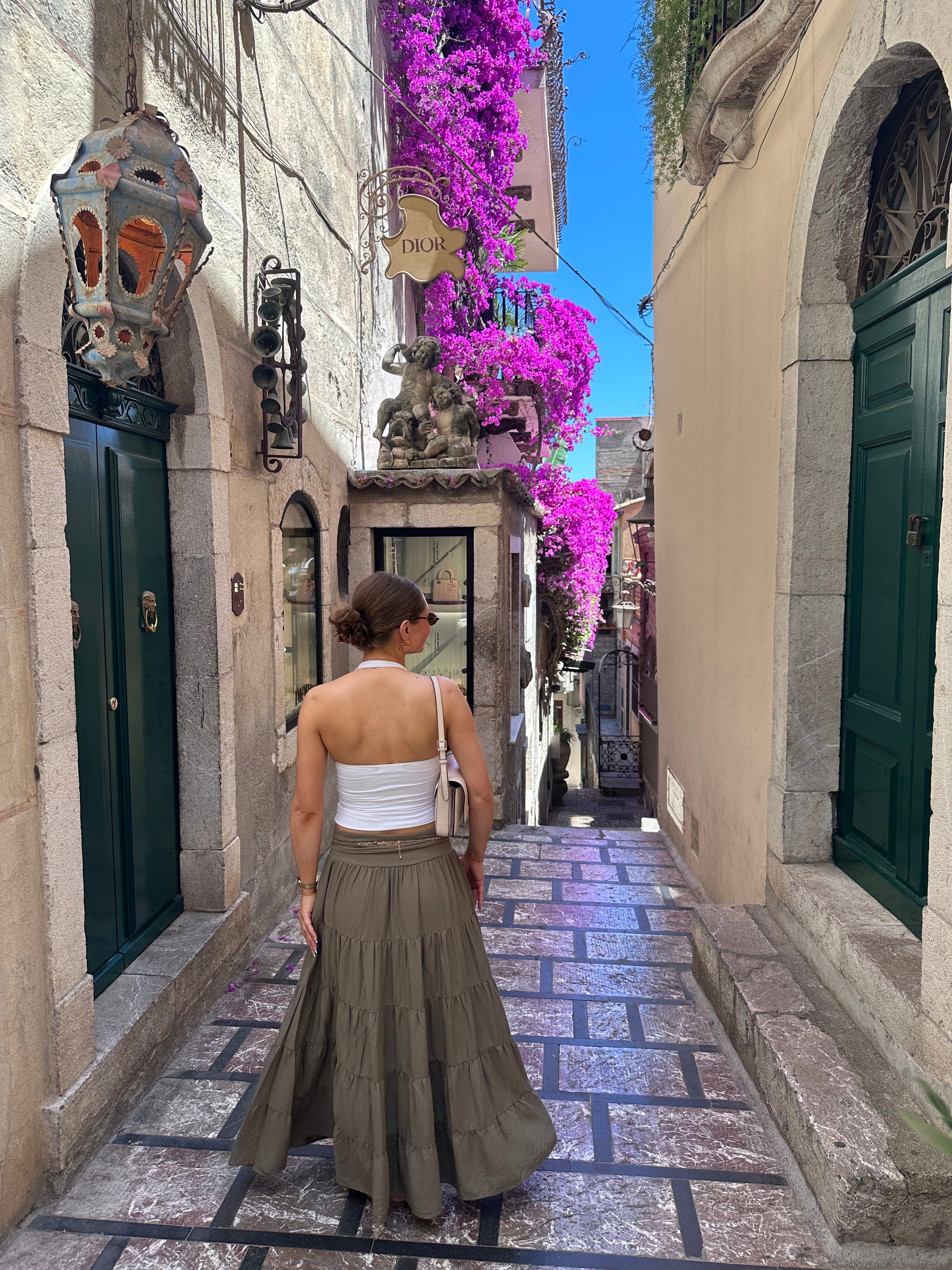 A woman in a white top and long skirt walking down a narrow street lined with flowers and stone buildings.