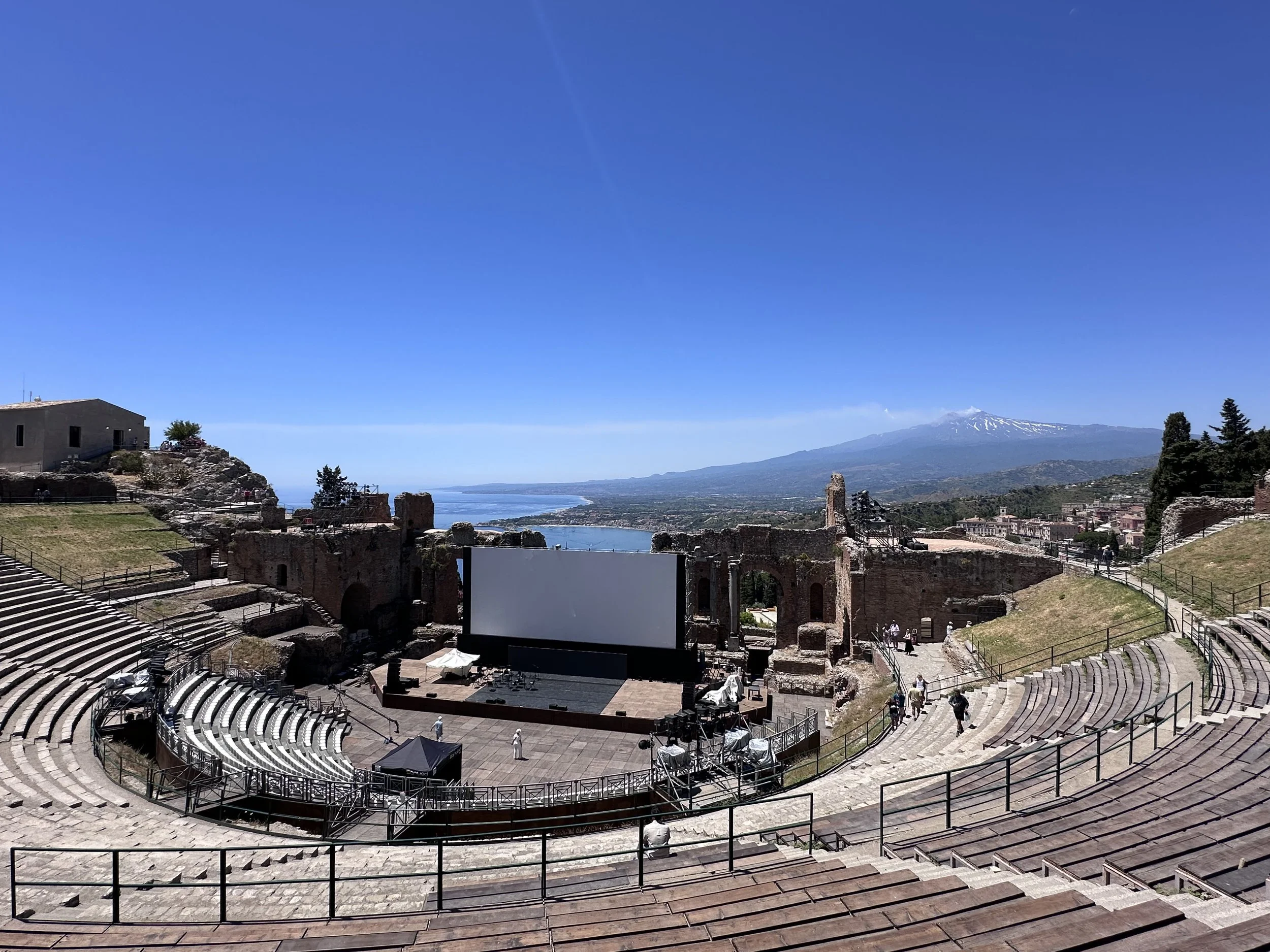 Ancient open-air amphitheater with a large screen, overlooking a coastline and a mountain with snow-capped peaks under a clear blue sky.