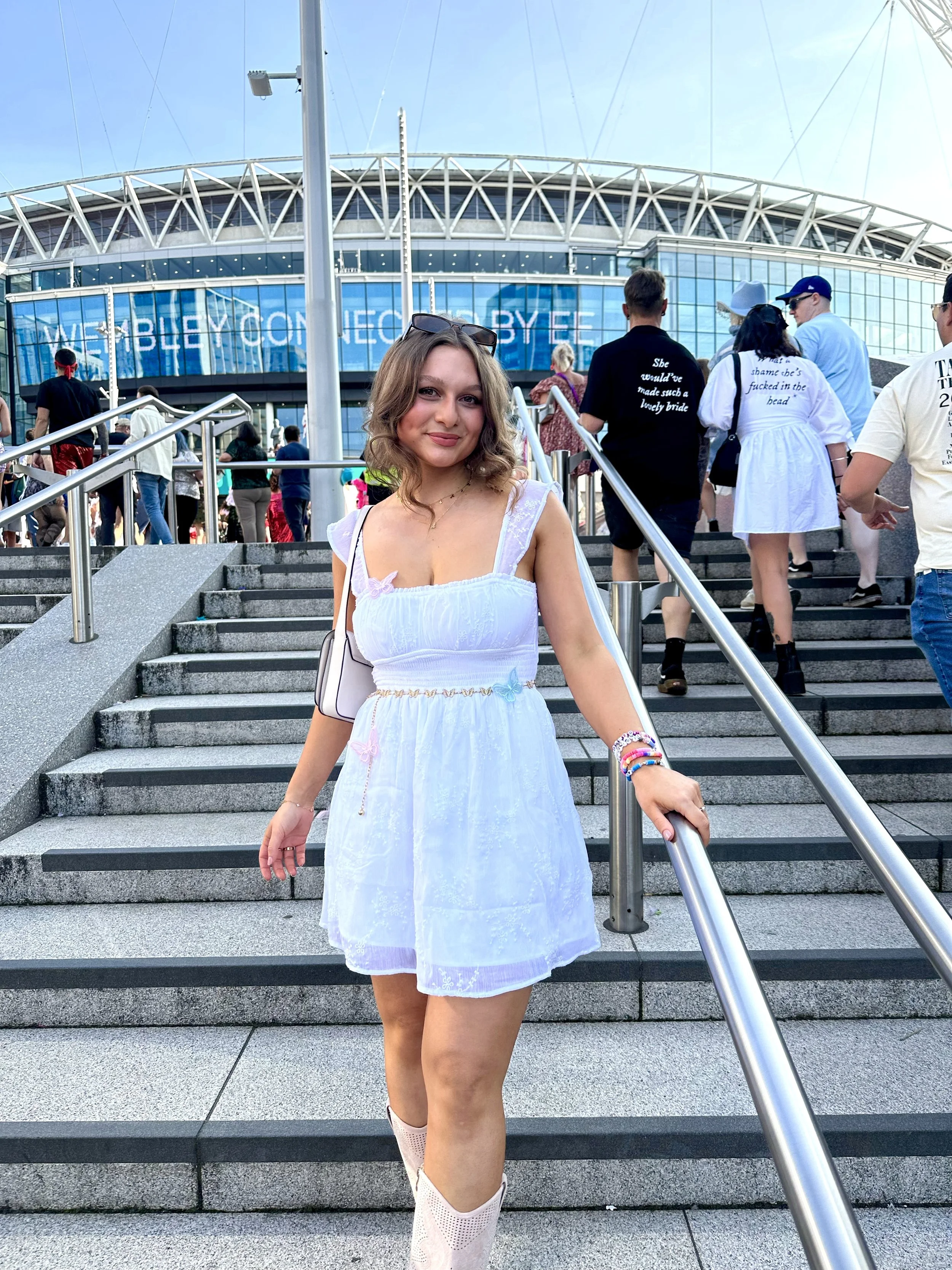 A woman posing in front of Wembley, wearing a white mini dress and pink cowboy boots at the Eras Tour