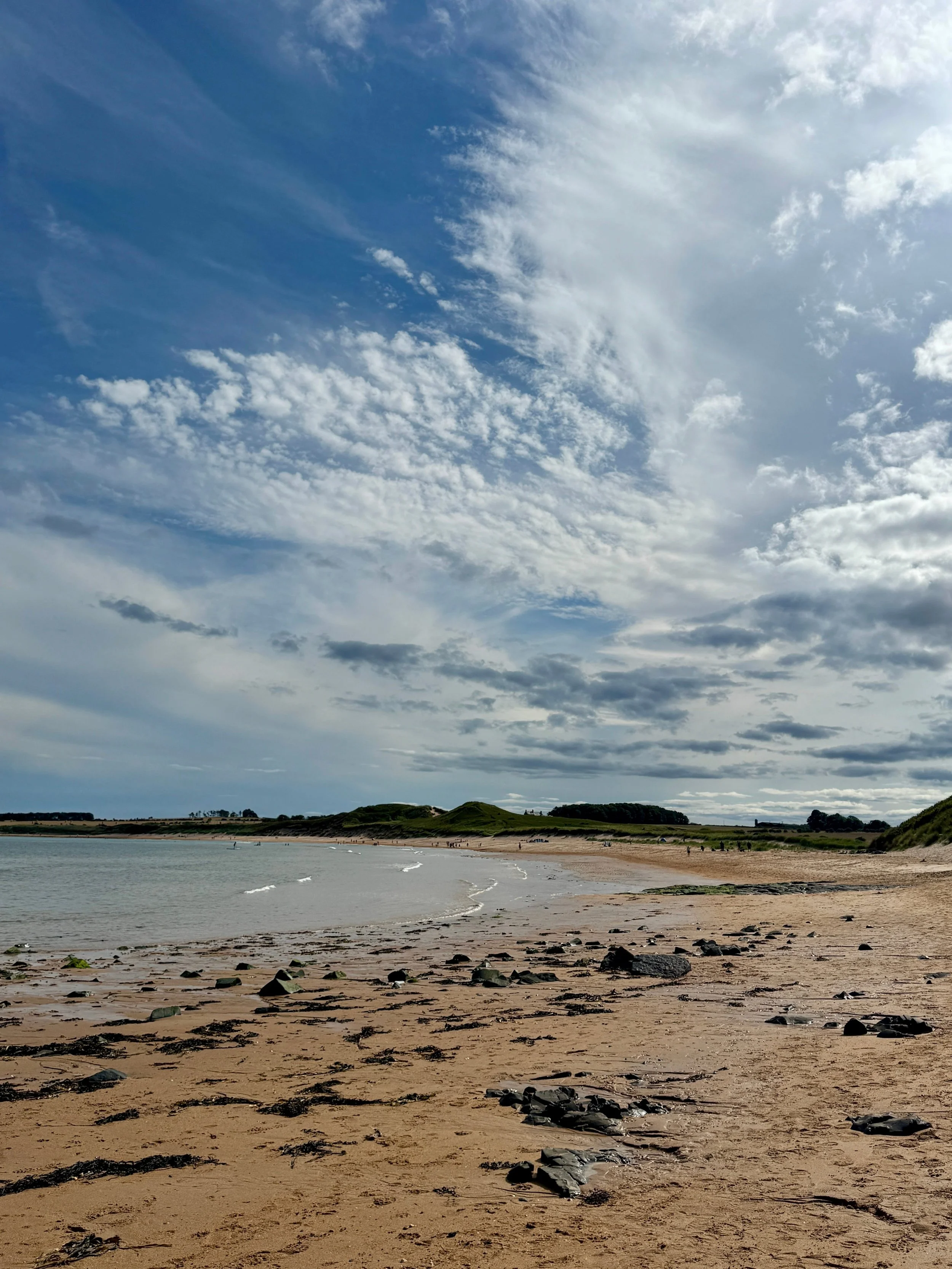 A quiet sandy beach with scattered dark rocks, gentle waves, and low green dunes beneath a vast blue sky streaked with clouds.