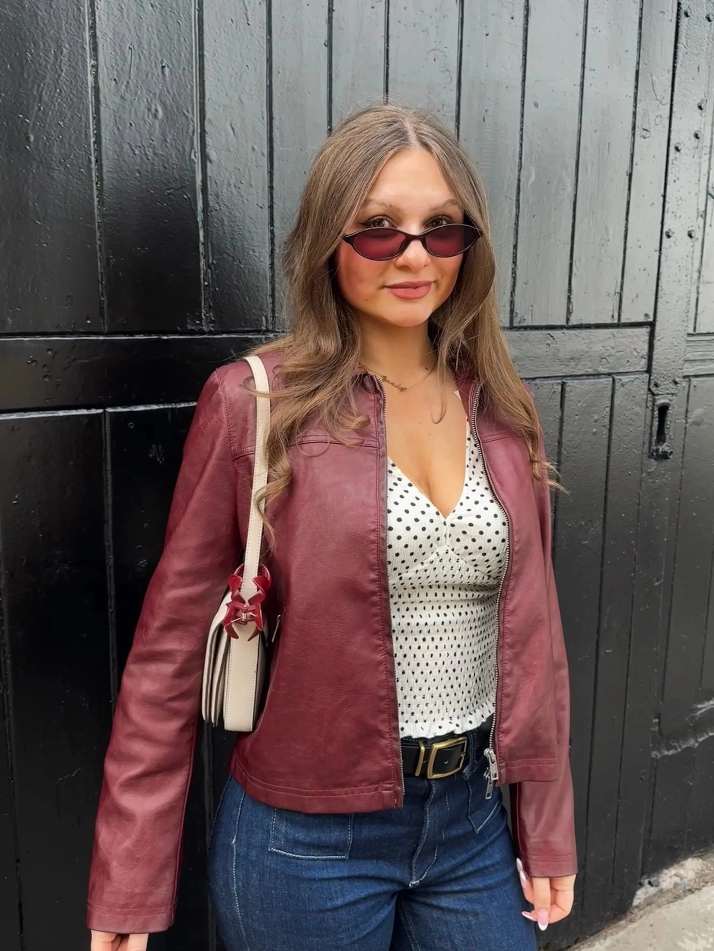 A woman in a maroon leather jacket and polka-dot top stands confidently against a black wooden wall. She wears sunglasses and carries a shoulder bag.