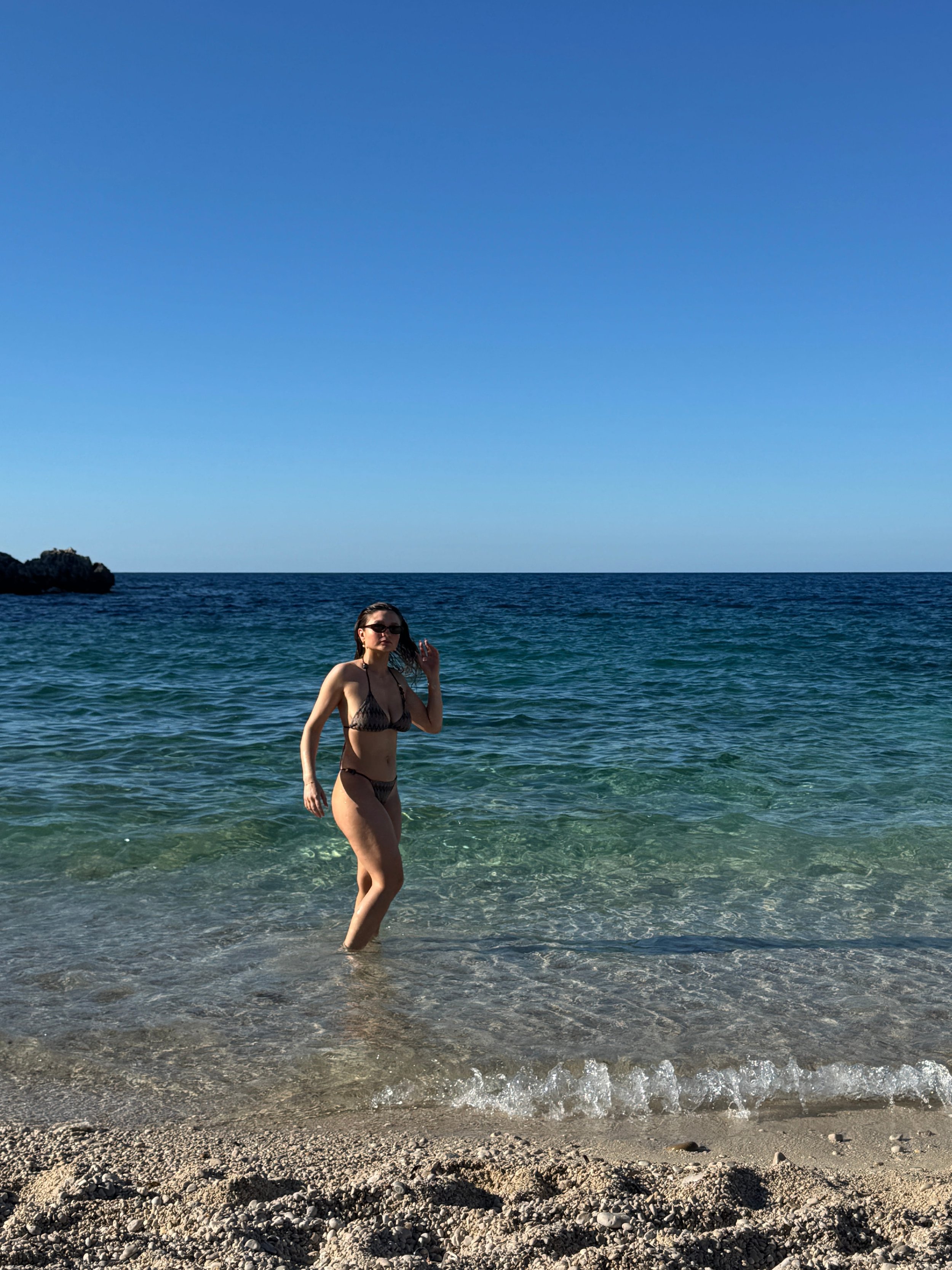A woman in a bikini strolls along the shoreline, waves gently lapping at her feet in the sparkling ocean.