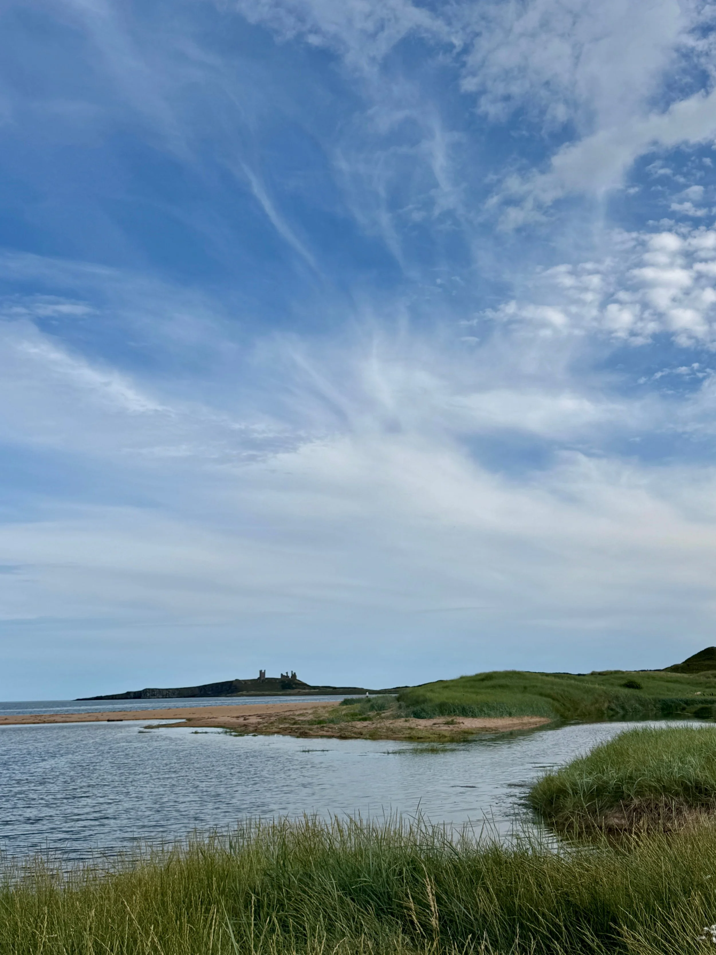 Serene coastal marsh with calm water, grassy dunes, distant shoreline, and a vast blue sky filled with wispy clouds.