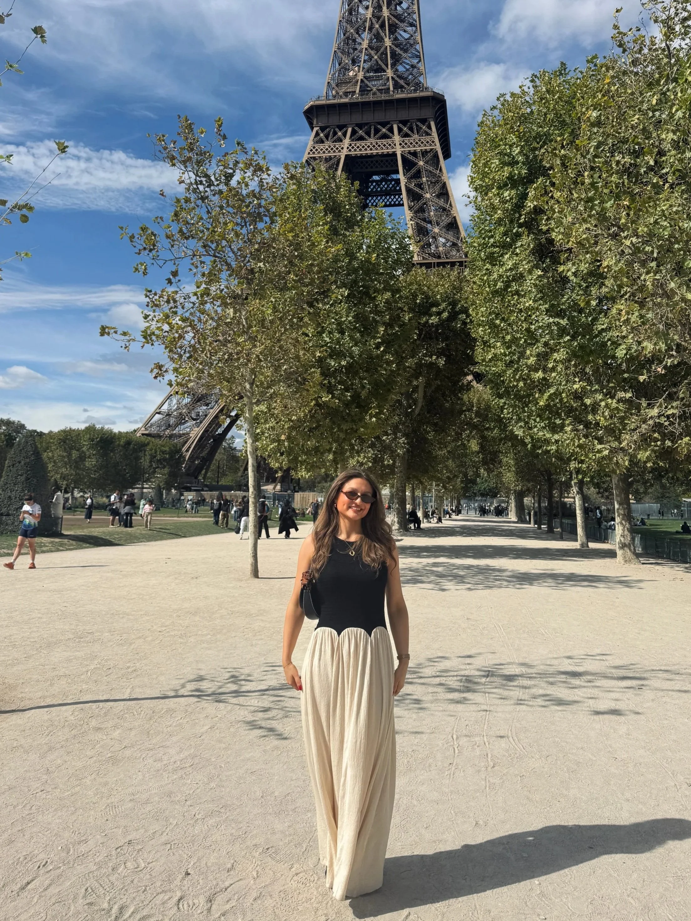 A woman in sunglasses, a black top, and flowing cream skirt stands beneath the Eiffel Tower in a sunny Paris park, framed by trees.