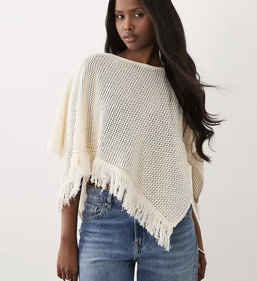 Woman wearing a cream-colored, knit poncho with fringe edges paired with high-waisted blue jeans, against a plain background.