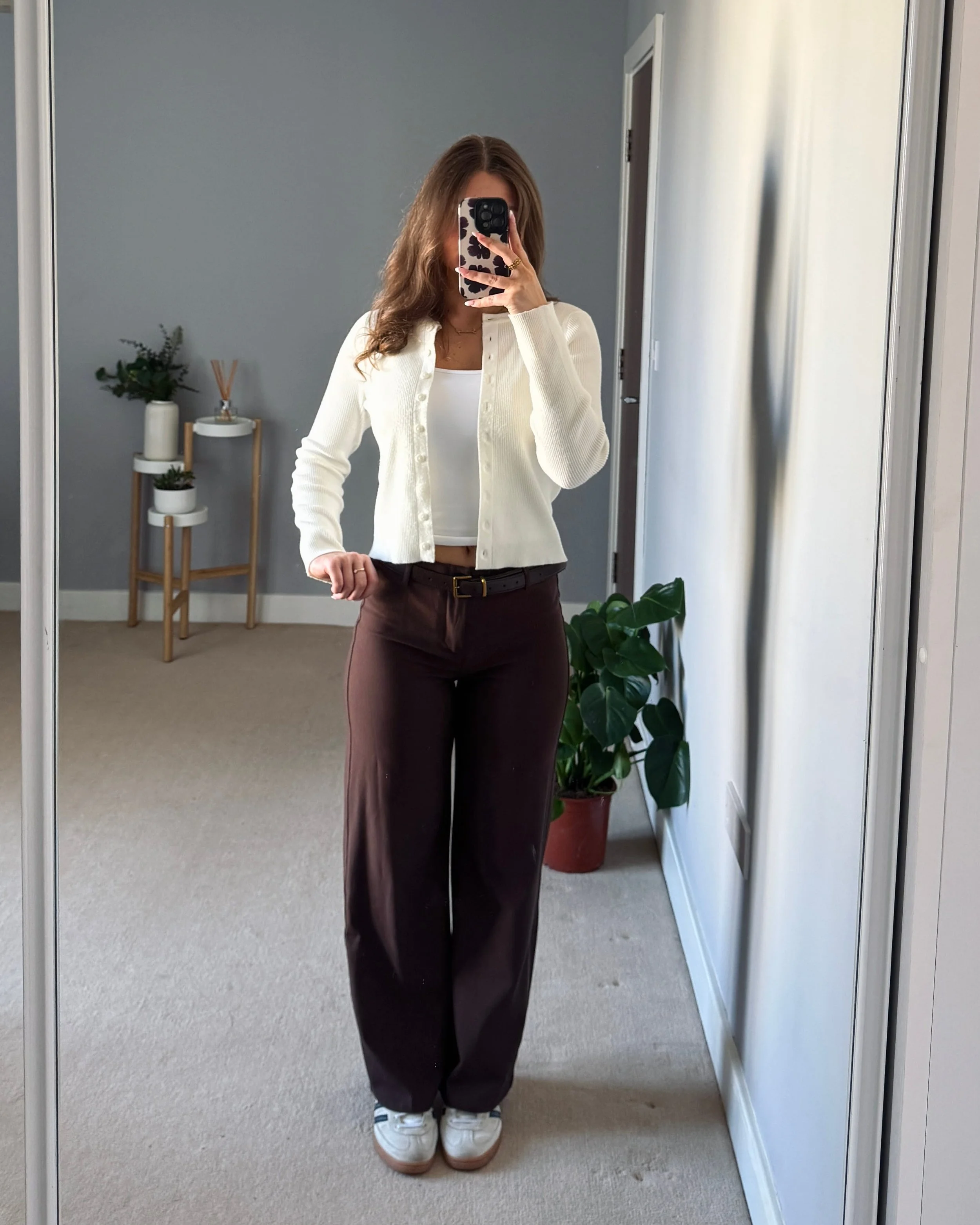 Woman taking a mirror selfie in a minimalist room. She wears a white cardigan, brown trousers, and trainers. There’s a potted plant beside her.