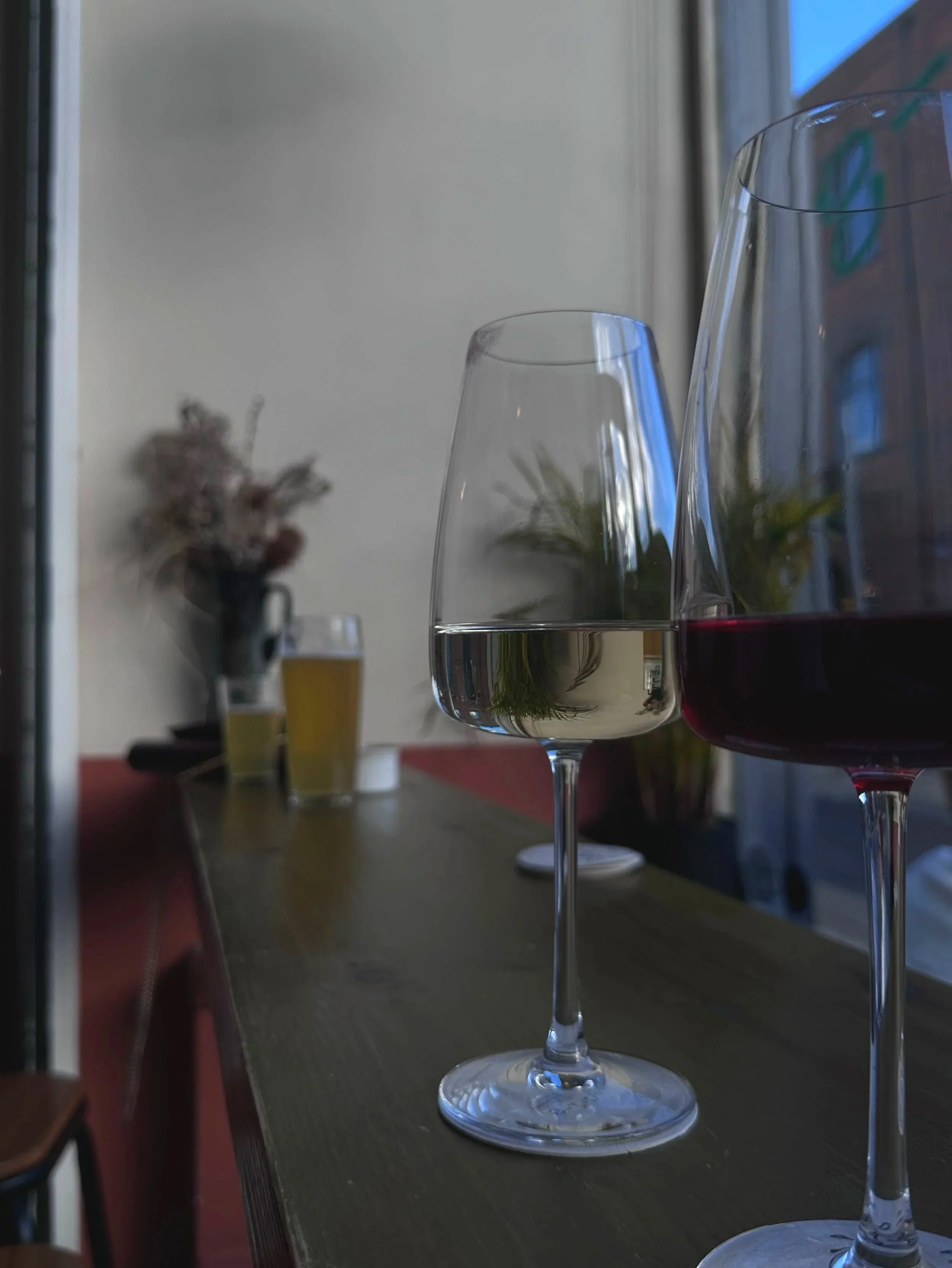 Two wine glasses, one with red and one with white wine, sit on a wooden bar by a window. A beer glass and dried flowers are in the background.