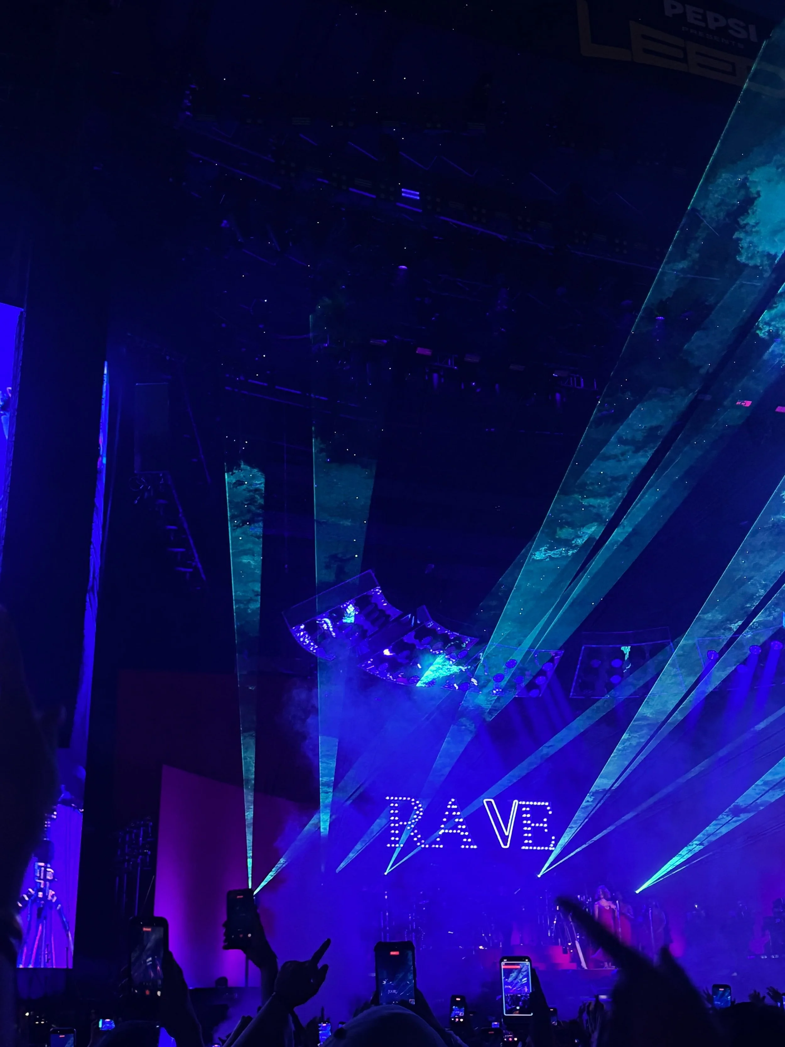A concert crowd raises phones beneath vivid blue and purple laser beams, with RAYE on stage and a “RAVE” sign creating an energetic nightclub atmosphere.