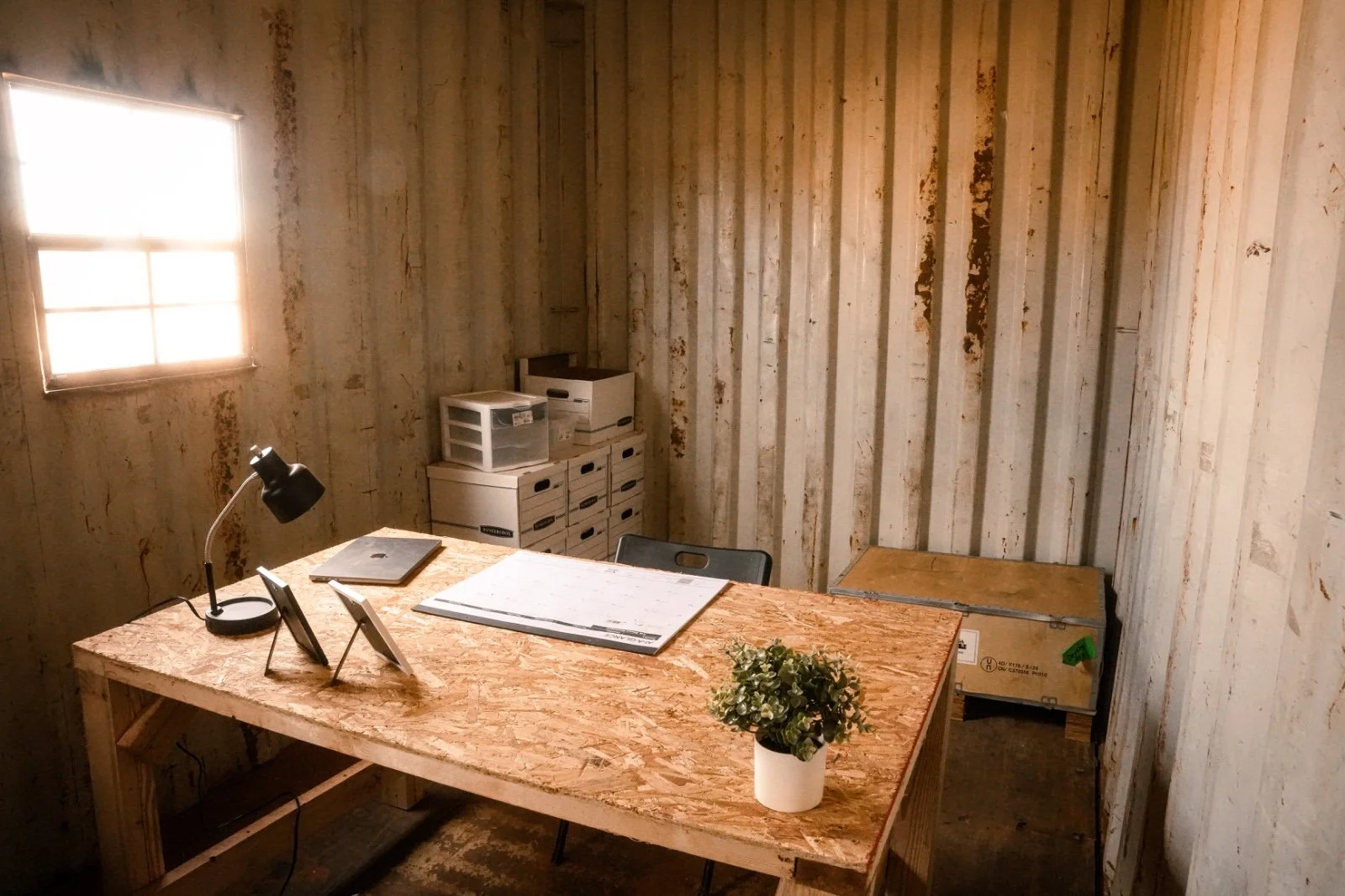 Ein kleines Büro mit einem Holzschreibtisch, auf dem ein Laptop, ein Tablet, eine Schreibtischlampe und eine Pflanze stehen. Im Hintergrund sind mehrere Aufbewahrungsboxen und eine Holzwand mit abblätternder Farbe.