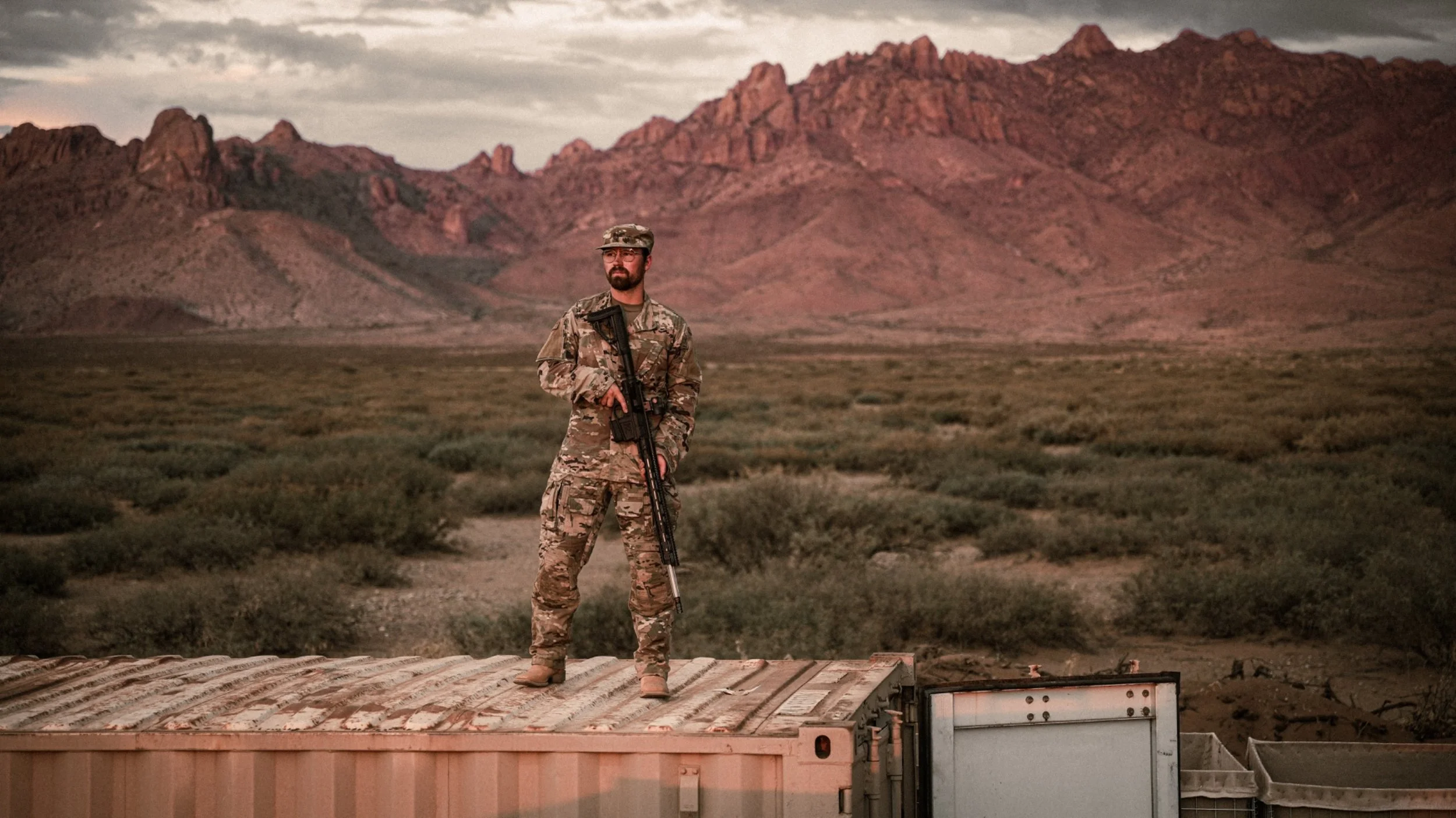 Ein Mann in Militäruniform steht auf einem großen Container in einer Wüstenlandschaft mit Bergen im Hintergrund.