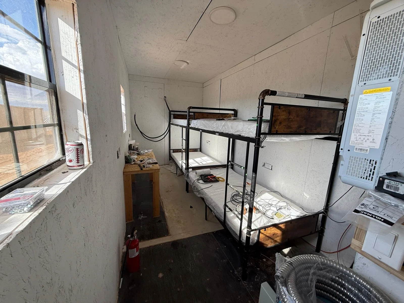 Ein unvollendetes Zimmer mit einem Metalbettgestell, Kabeln, einem kleinen Holzschreibtisch, einer Fensterfront und einem Heizgerät.