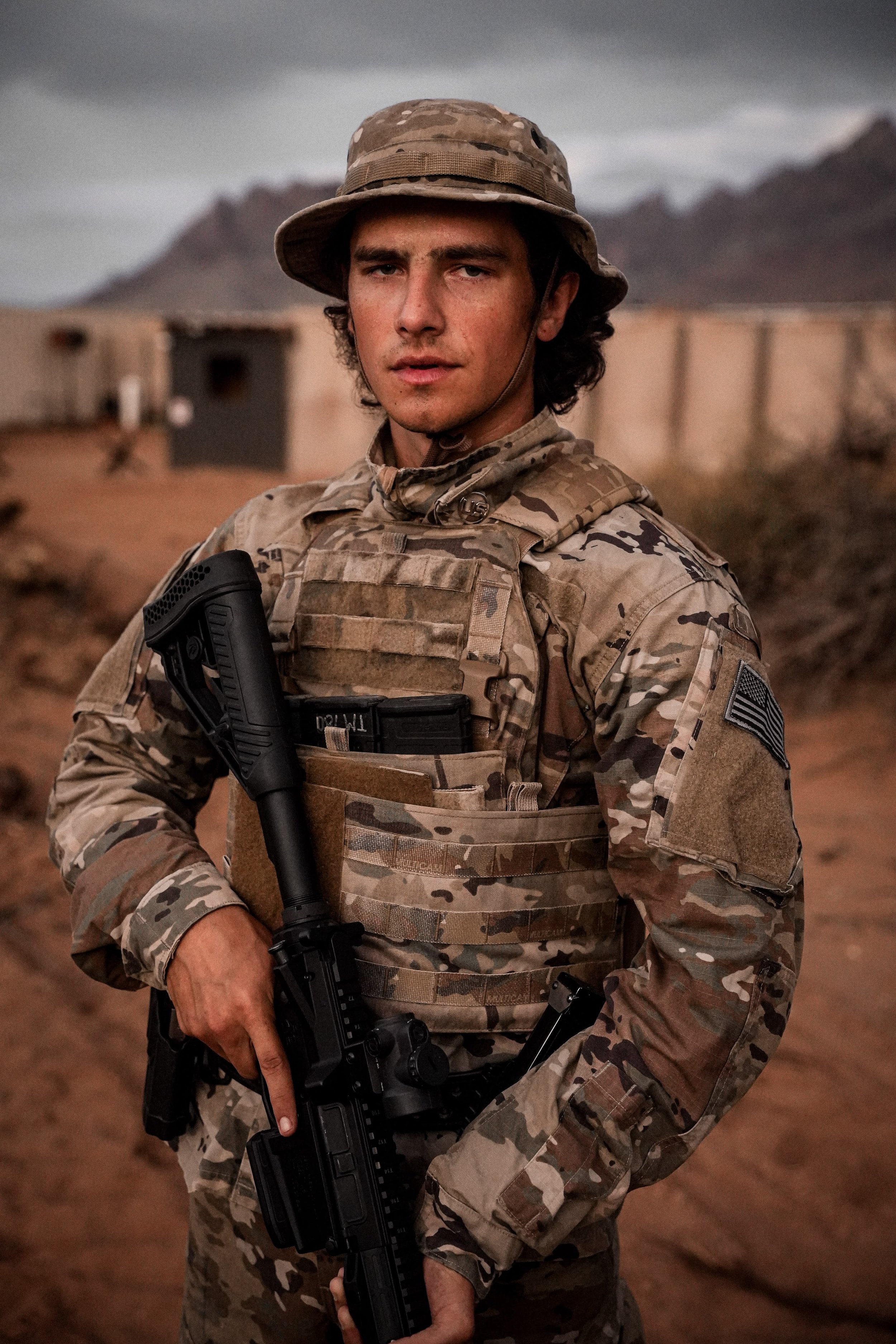 Ein Soldat in Camouflage-Uniform steht in einer Wüstenlandschaft mit Bergen im Hintergrund, trägt einen Hut und hält eine Waffe.