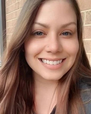 Close-up of a smiling young woman with light makeup and long brown hair, standing outdoors in front of a brick wall.