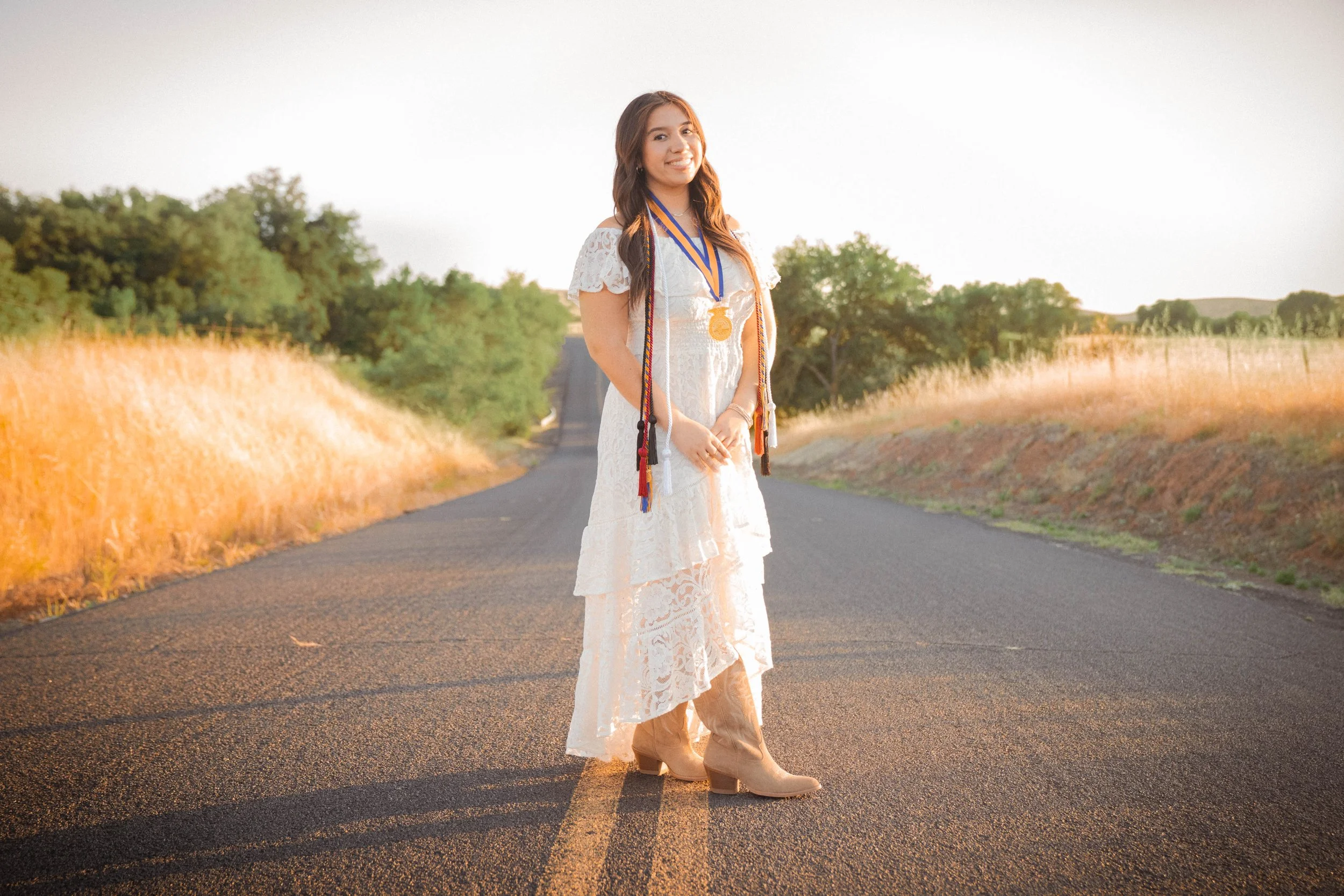 A young woman standing on a rural road at sunset, wearing a white lace dress, tan cowboy boots, and a medal around her neck. She is smiling and looking at the camera, with fields and trees in the background.