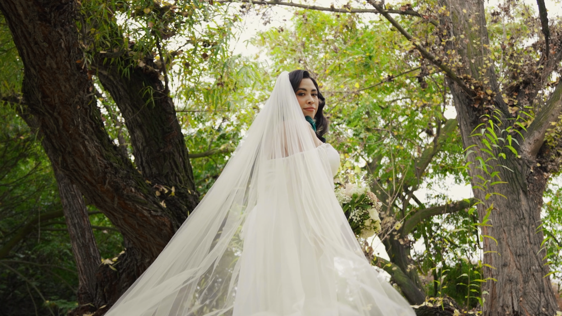A bride in a white wedding gown with a long veil standing in a lush, green outdoor setting with large trees and foliage.