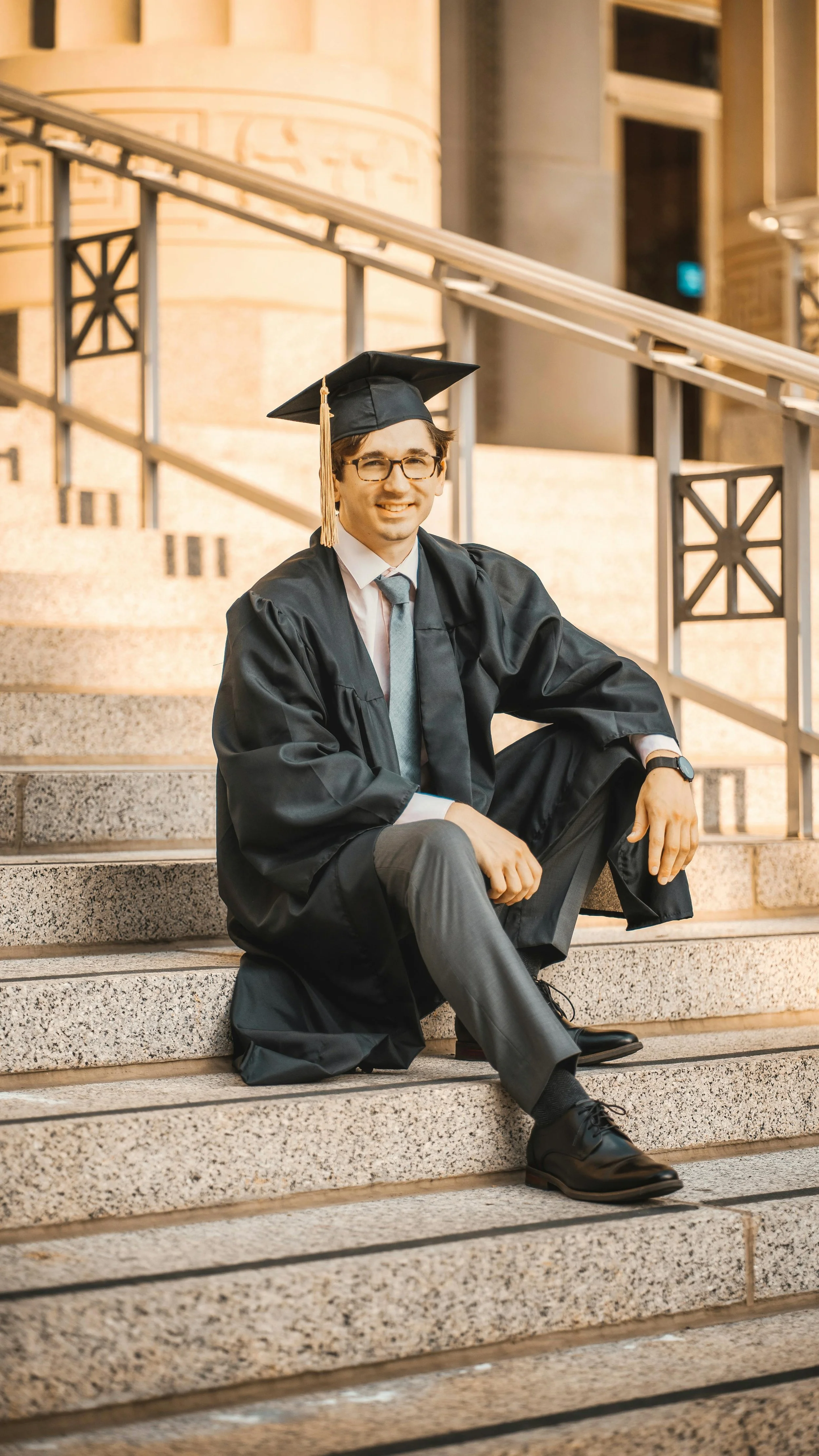 A young man in graduation cap and gown sitting on outdoor stone steps, smiling at the camera.