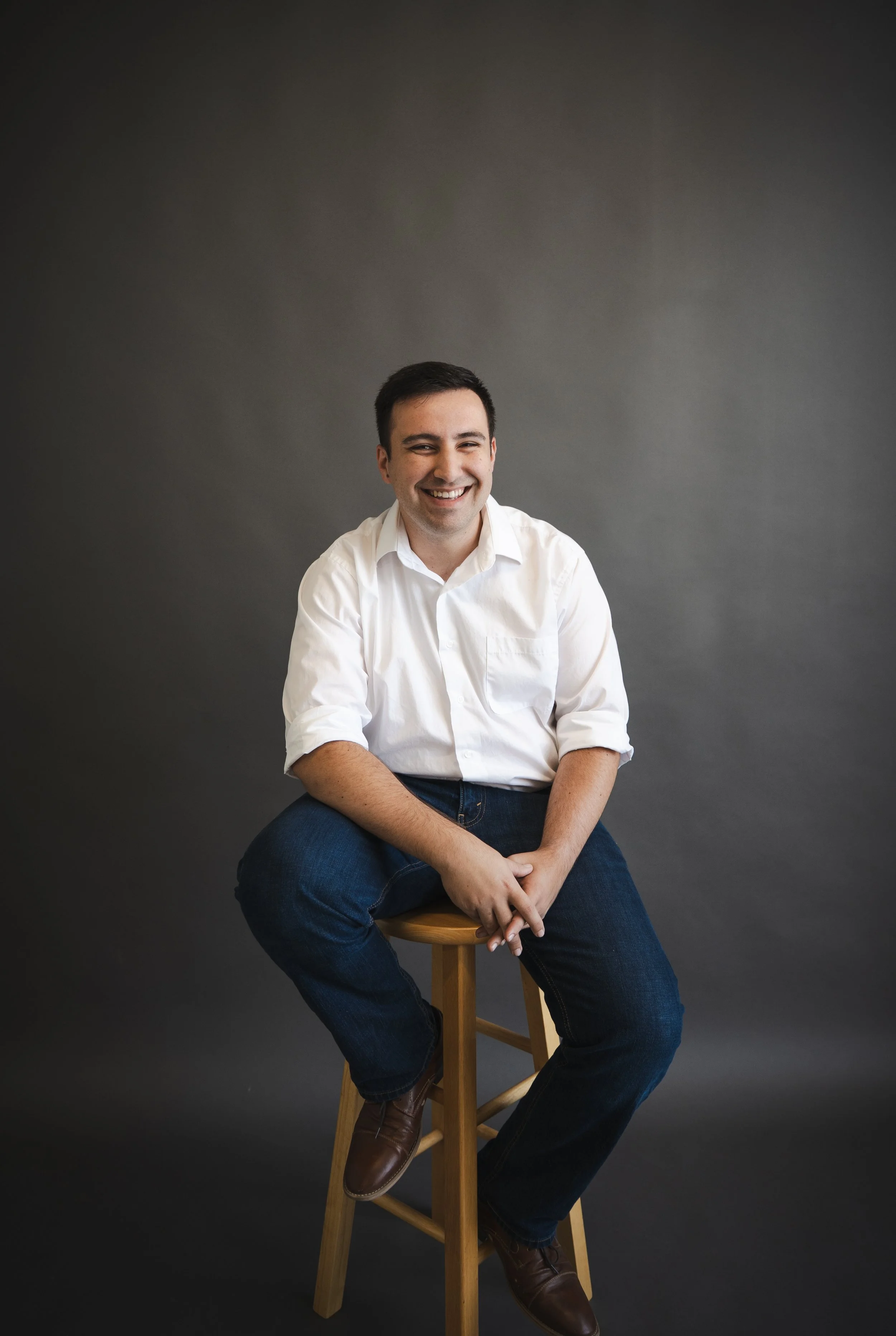 A smiling man in a white shirt and jeans sitting on a wooden stool against a dark gray background.
