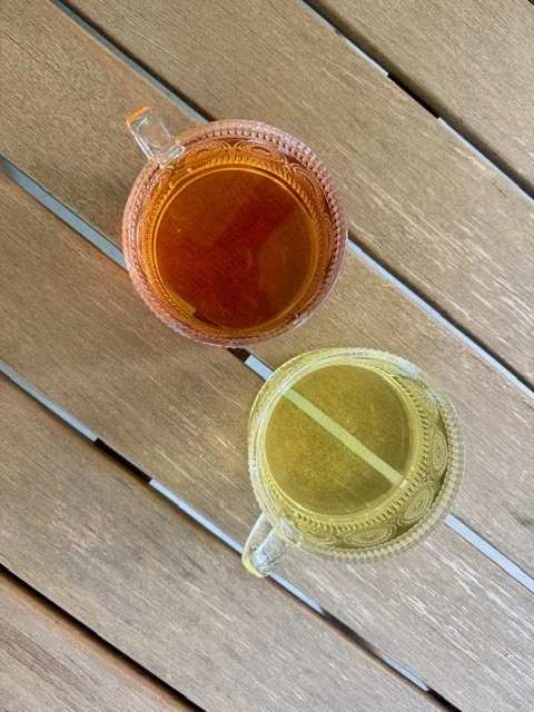 Two glass pitchers, one filled with tea or amber-colored beverage and the other with a light yellow drink, on a wooden outdoor table.