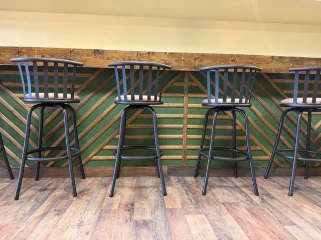 Four black barstools lined up in front of a wooden counter with a diagonal slat design on the front, against a green wall.