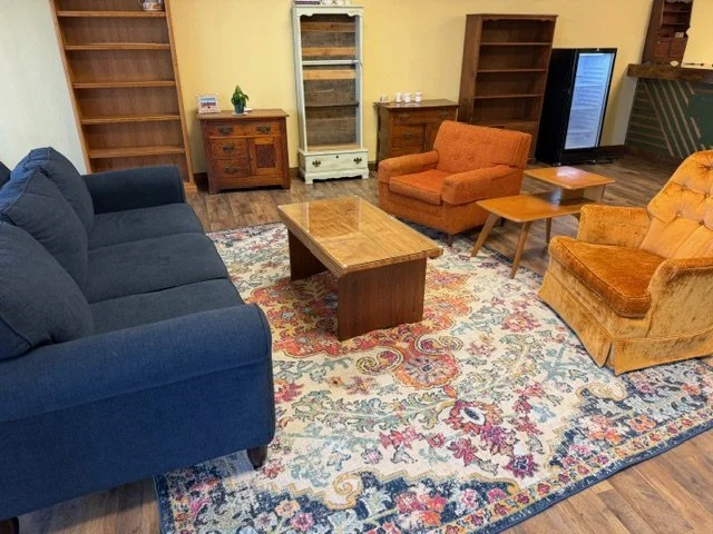 Living room with a blue sofa, two orange armchairs, a glass coffee table, wooden bookshelves and cabinets, a patterned area rug, and a wooden floor.