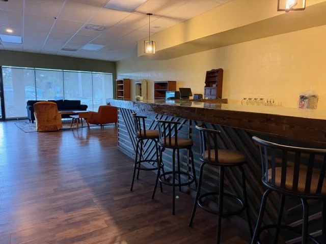 Empty bar area with high chairs, a wooden countertop, and shelves on the wall, with a seating lounge in the background near large windows.