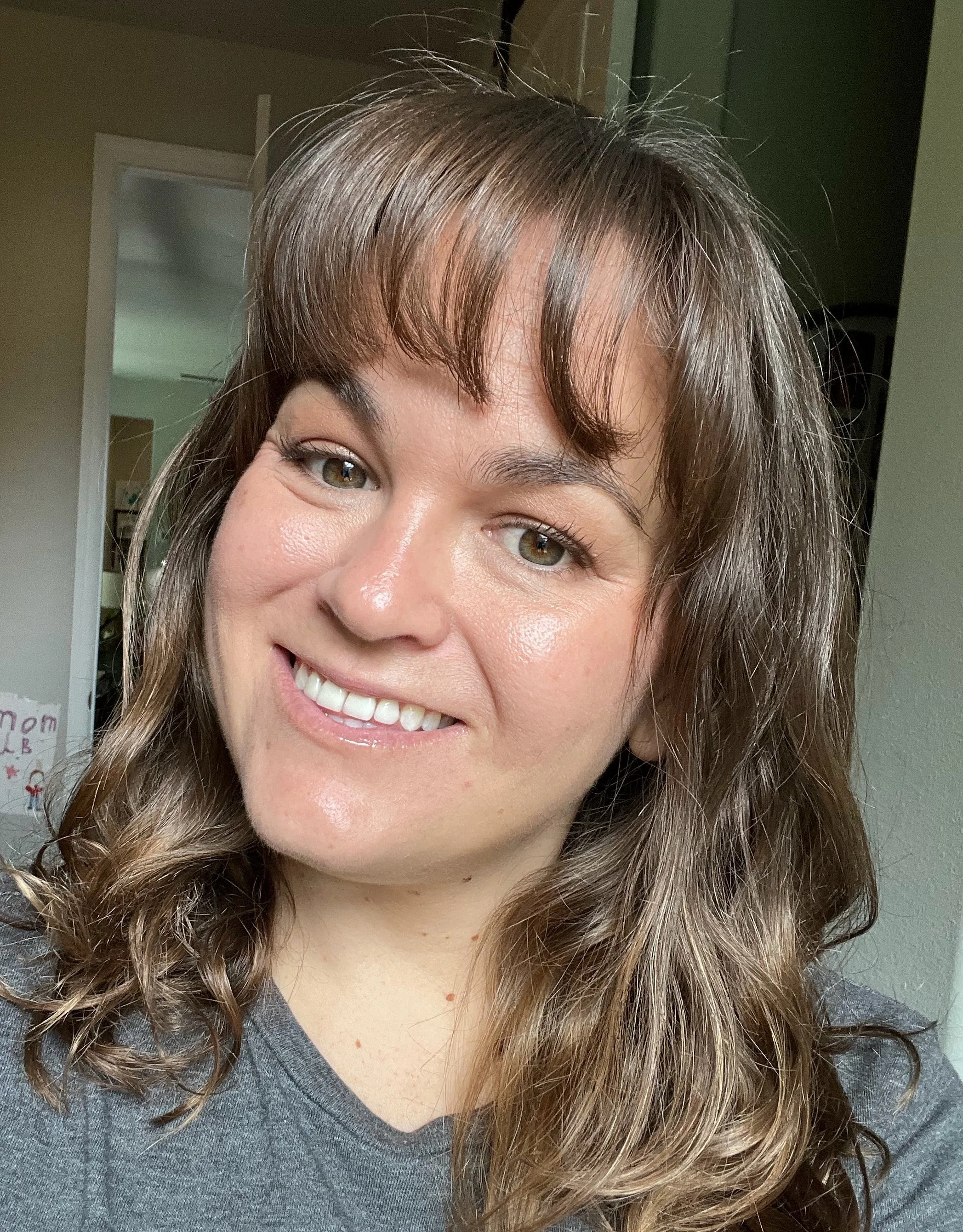 Close-up of a smiling woman with wavy brown hair, wearing a gray shirt, indoors.