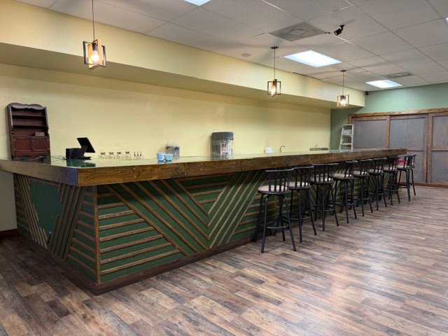Empty bar counter with black stools, wooden shelf, and hanging pendant lights in a warmly lit room with wood flooring and pastel green walls.