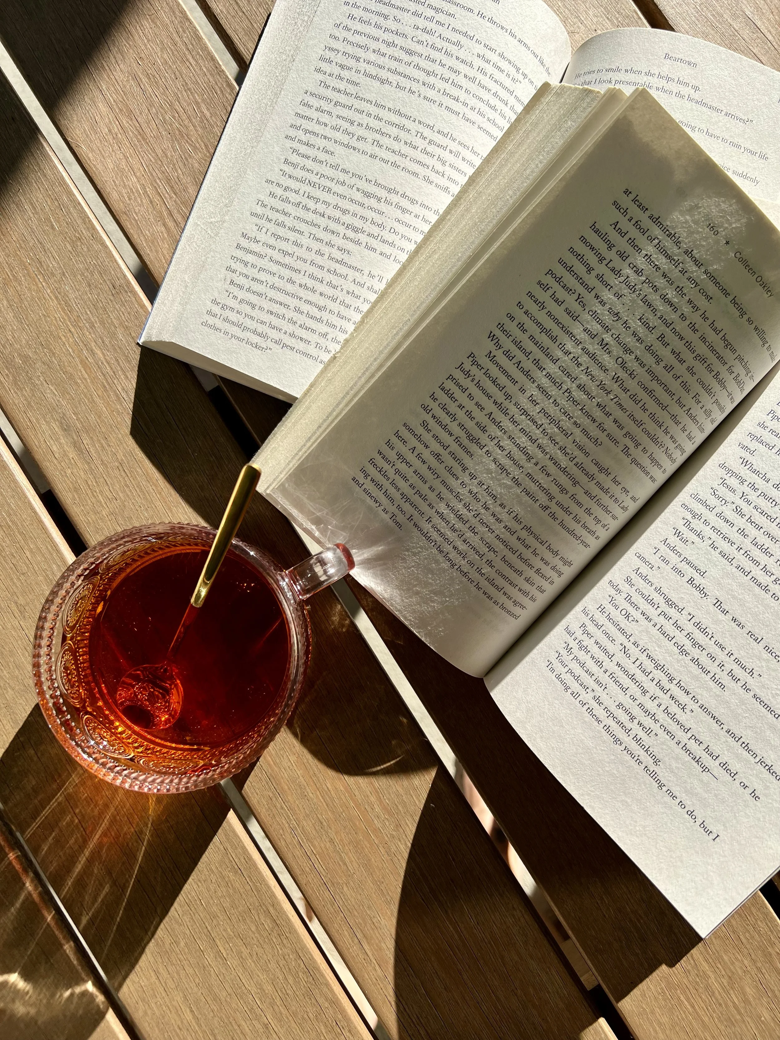 Open book, glass of dark red beverage with a straw, and a closed book on a wooden table.