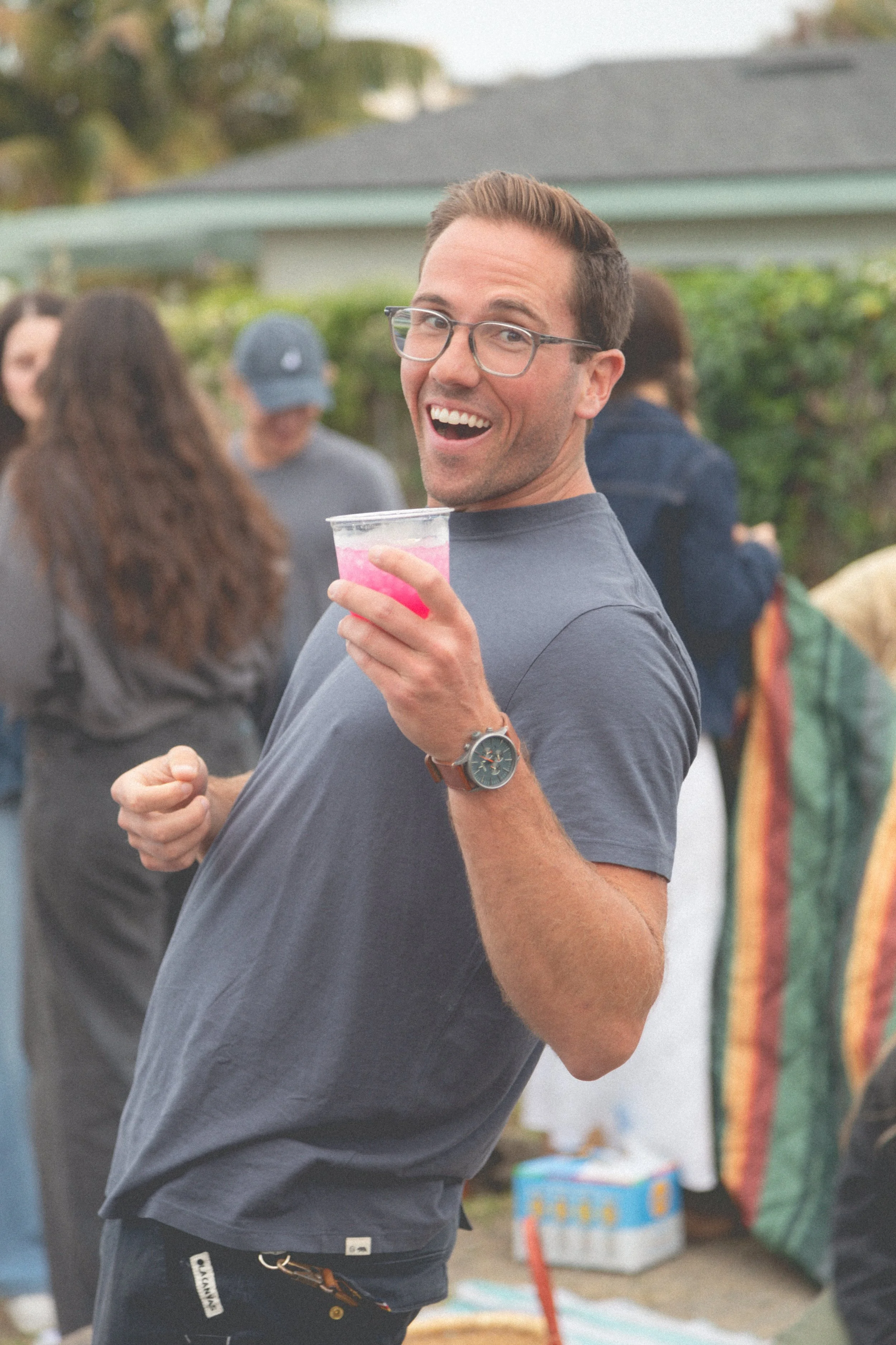 A man with glasses smiling and holding a pink drink at an outdoor gathering with other people in the background.