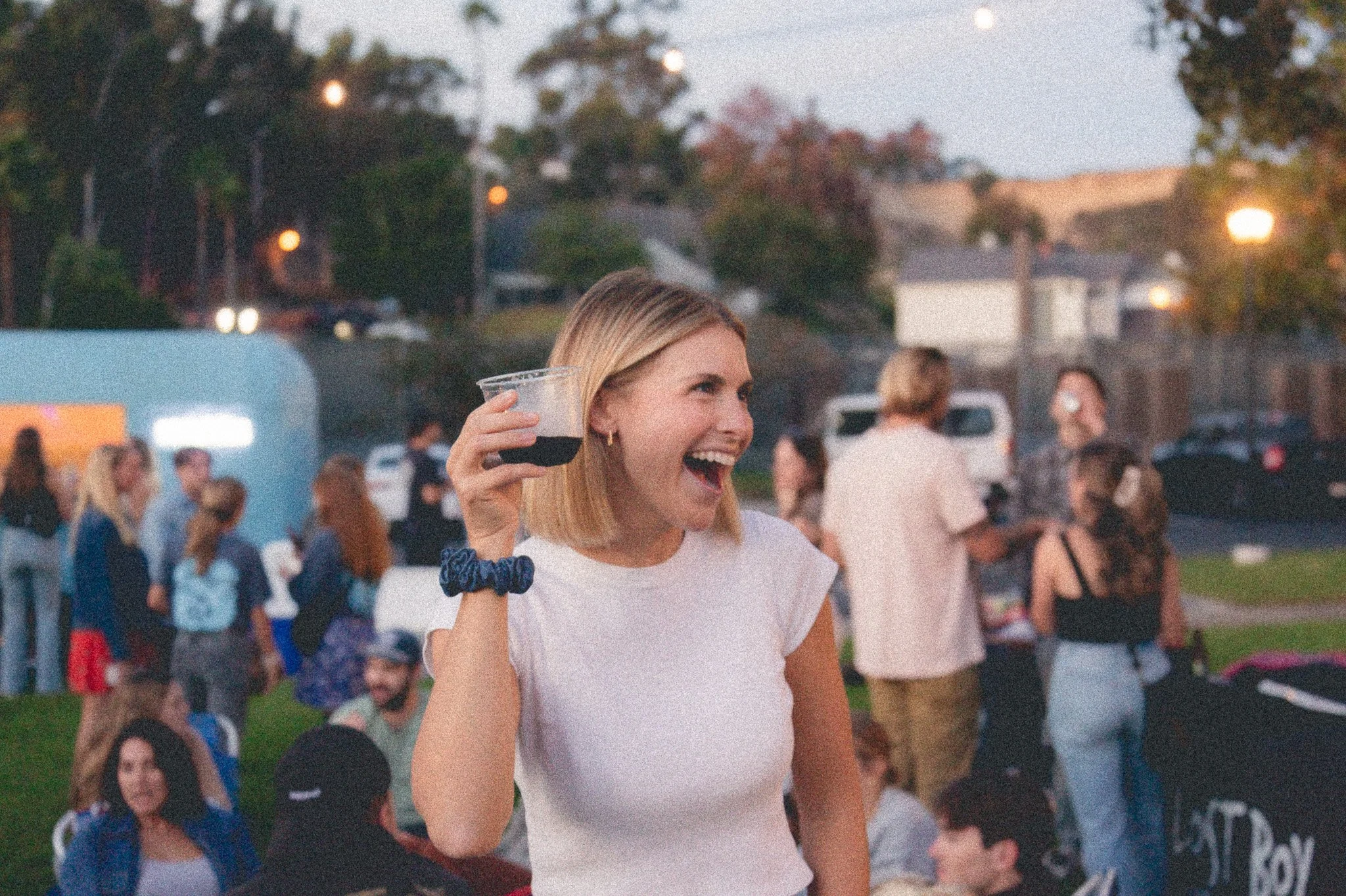 A woman smiling and holding up a drink at an outdoor gathering during dusk, with people socializing in the background.