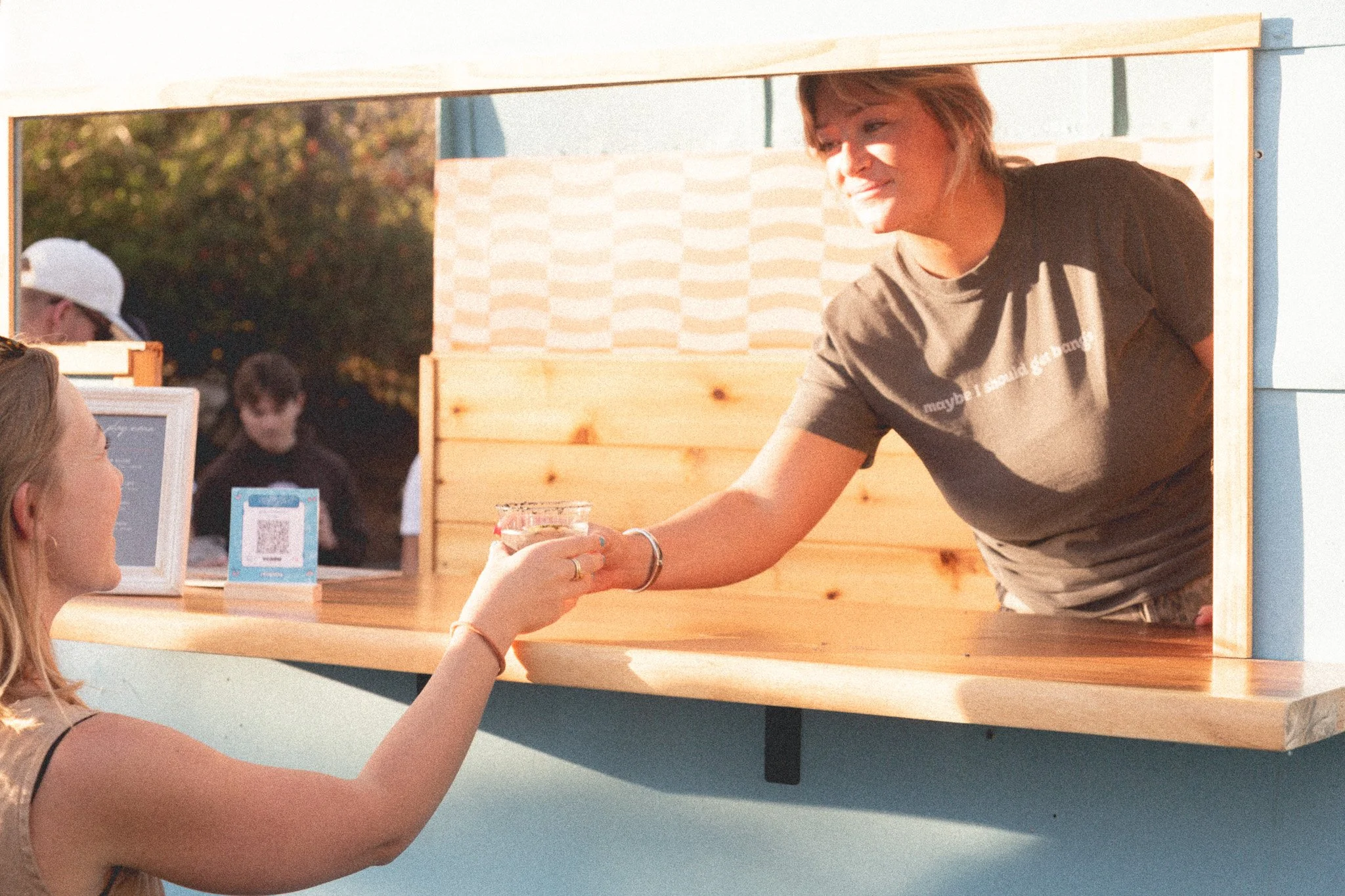 A woman serving a drink through a small window to a young woman at an outdoor booth, with other people in the background.